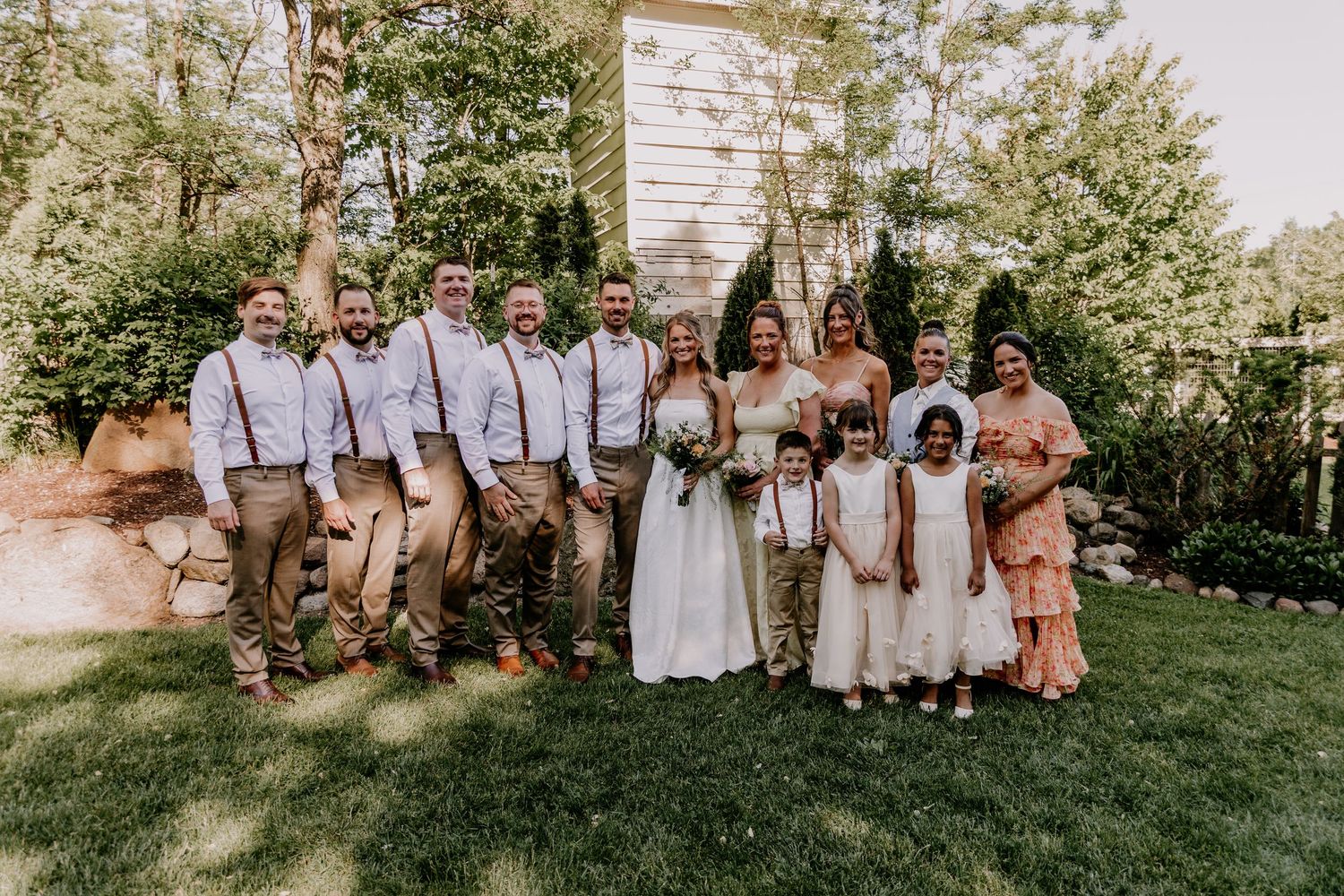 Wedding party poses outdoors in garden setting, groomsmen in suspenders, bride in white gown, flower girls, and guests.