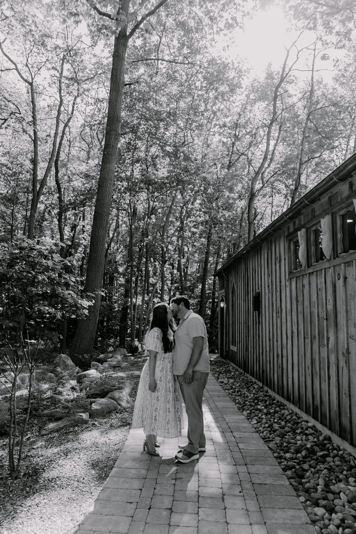 Black and white photo of a couple sharing a tender kiss on a brick pathway surrounded by lush trees.