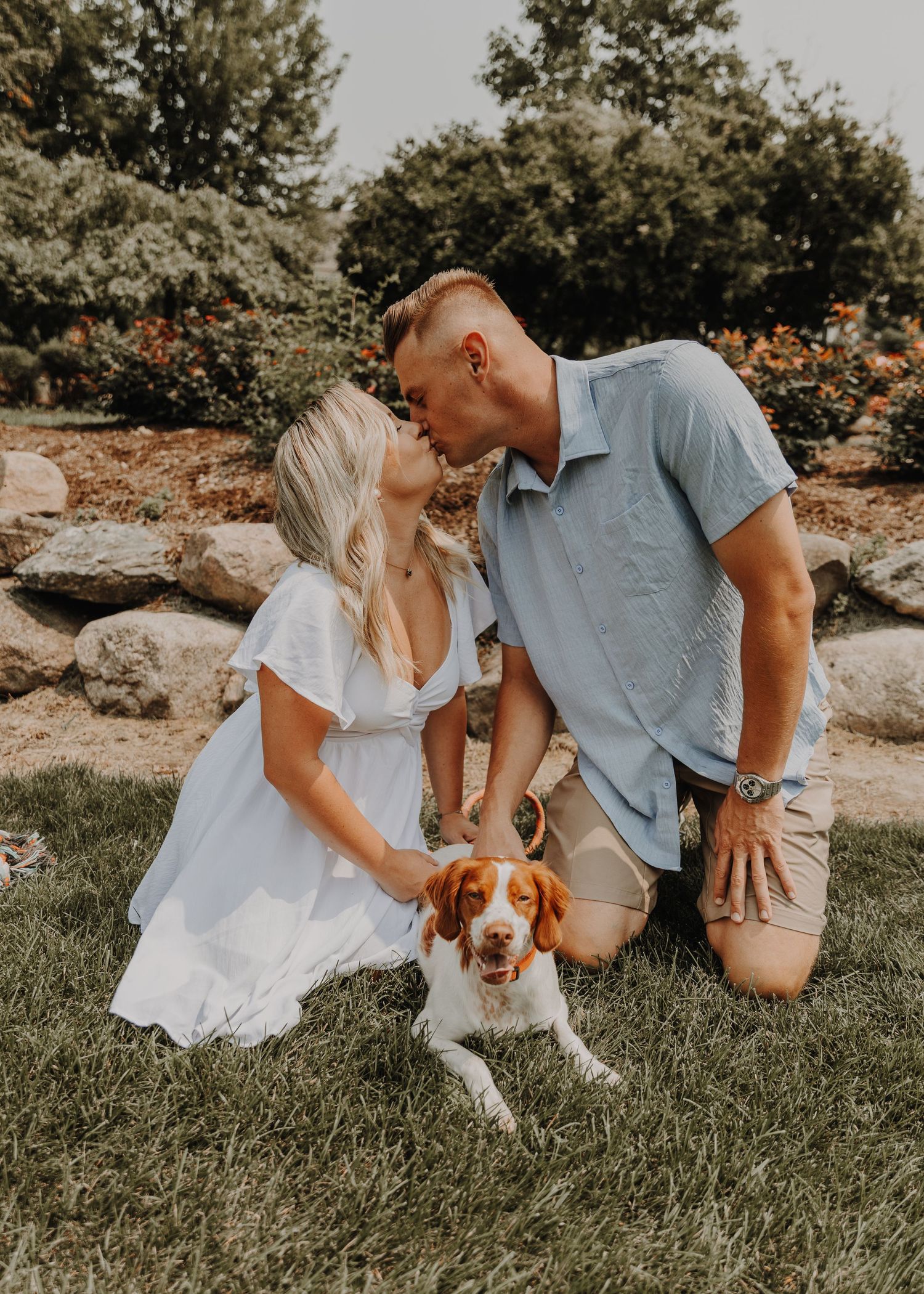 A couple in white and blue outfits plays with their beagle puppy on grassy ground during a sunny outdoor photoshoot.
