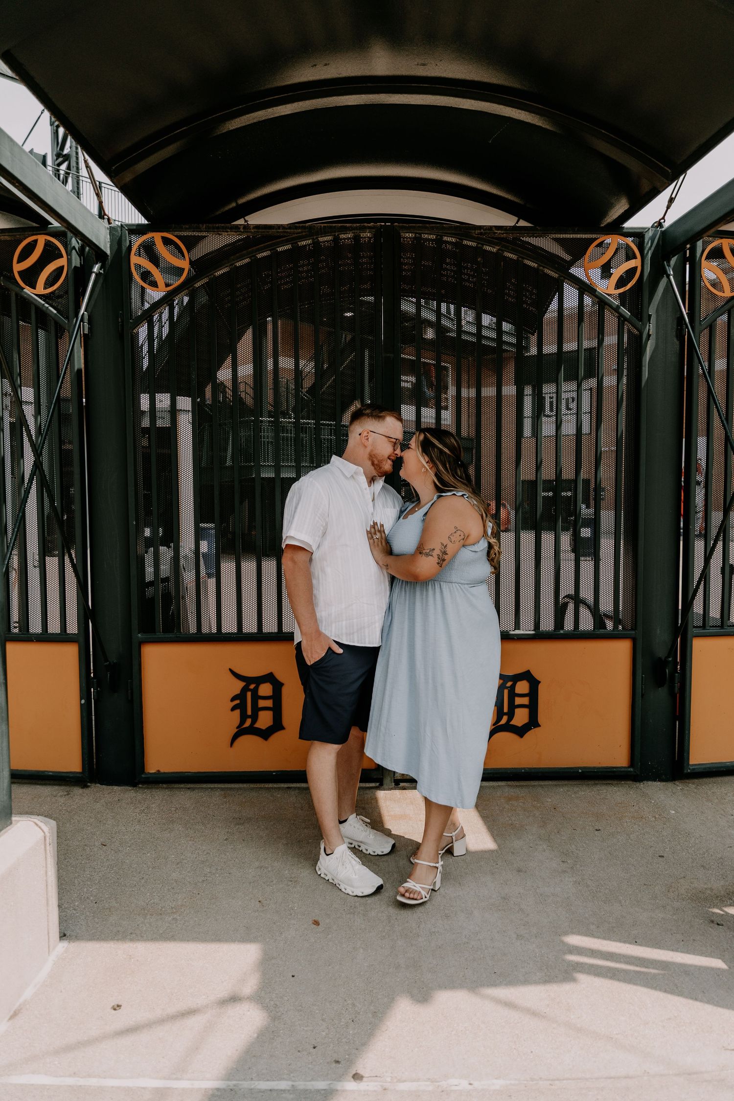A couple shares an intimate moment at the entrance of a stadium with orange and black architectural details.