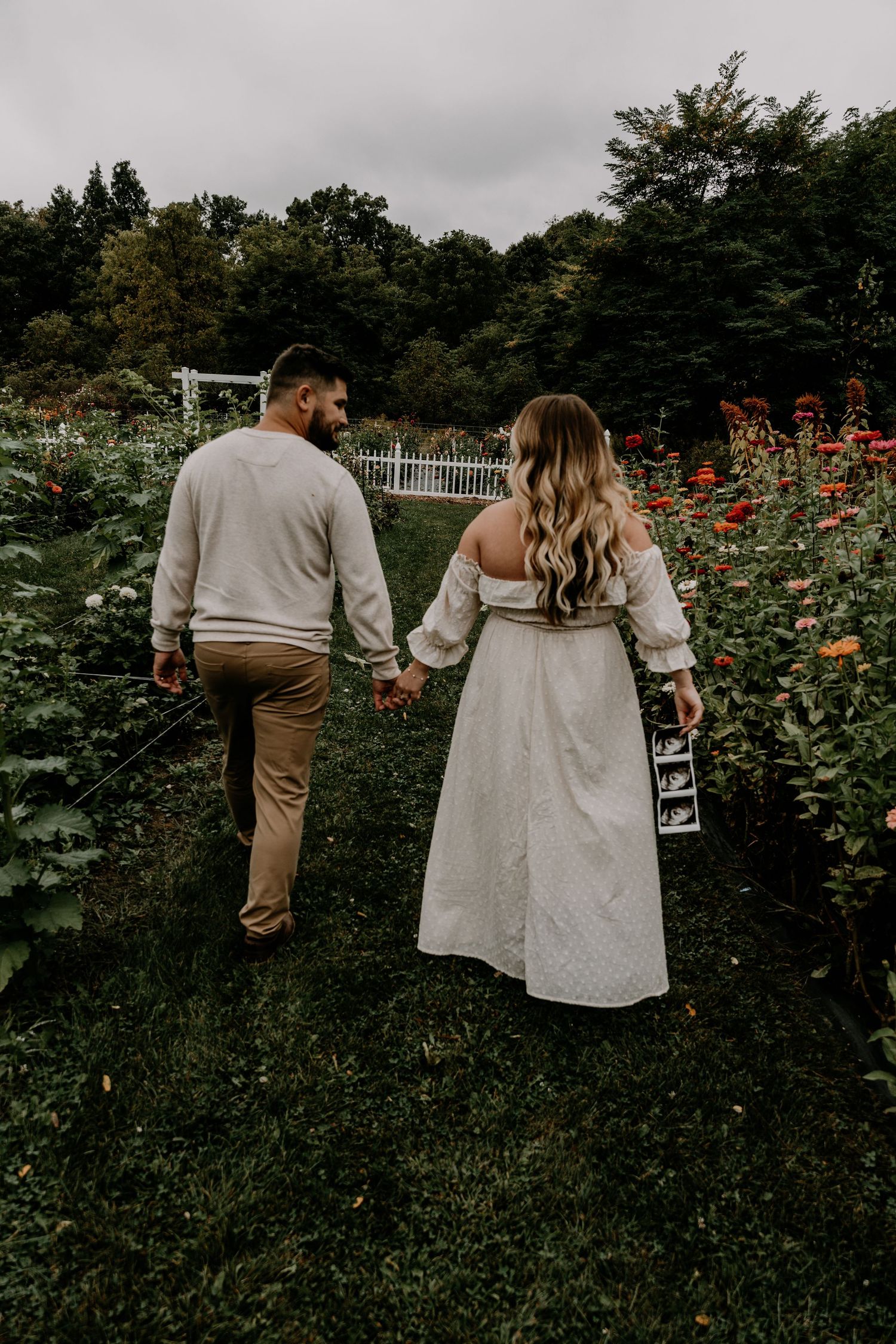 Couple walking through garden pathway with white picket fence and flowers.