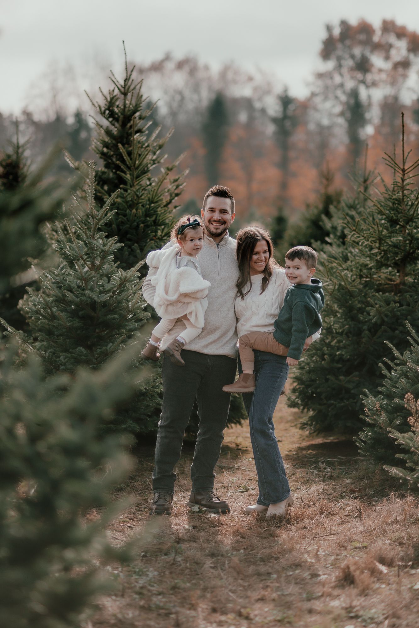 Family of four posing together among evergreen trees at a Christmas tree farm.