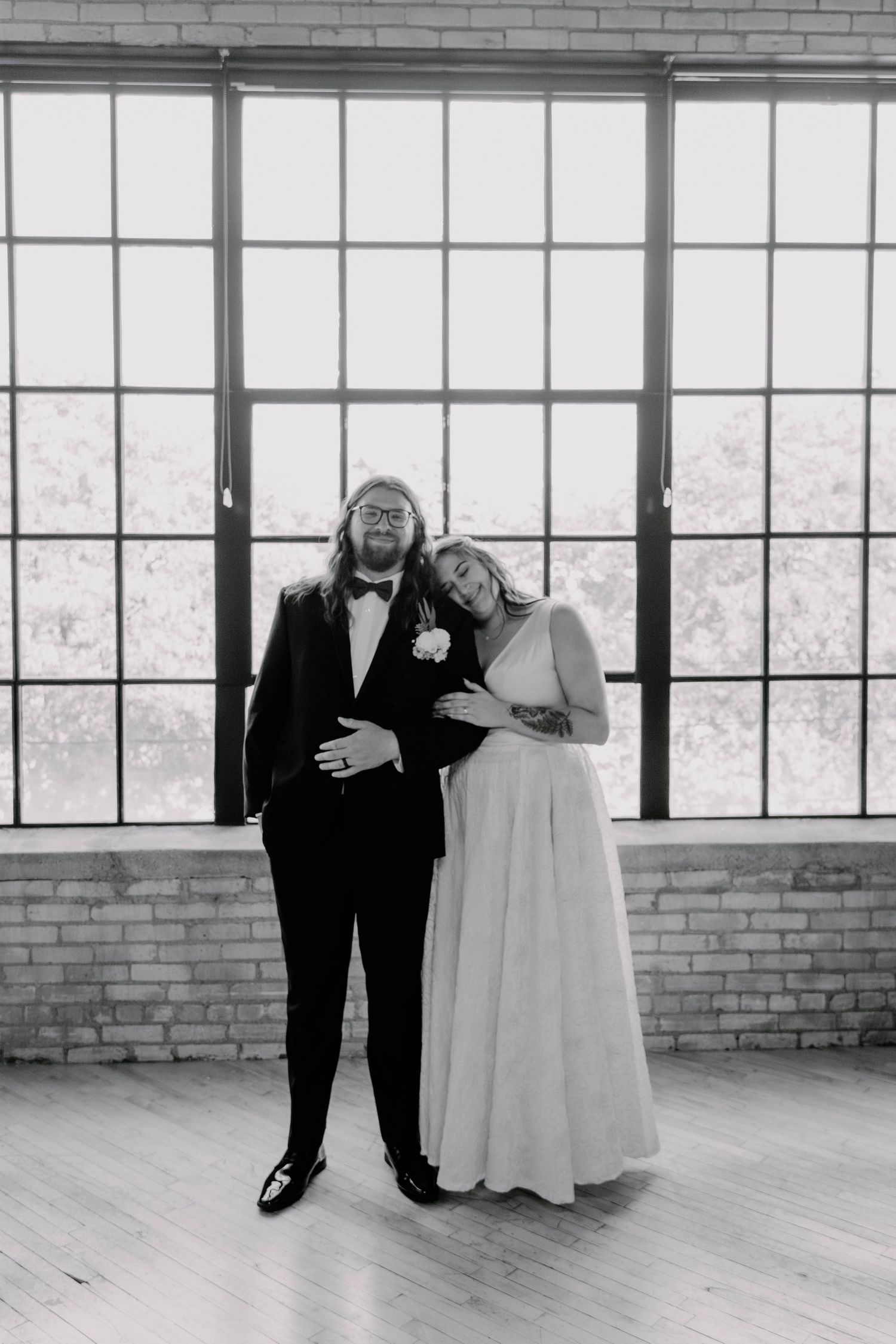 A couple in formal wedding attire shares an intimate moment in front of large industrial windows.