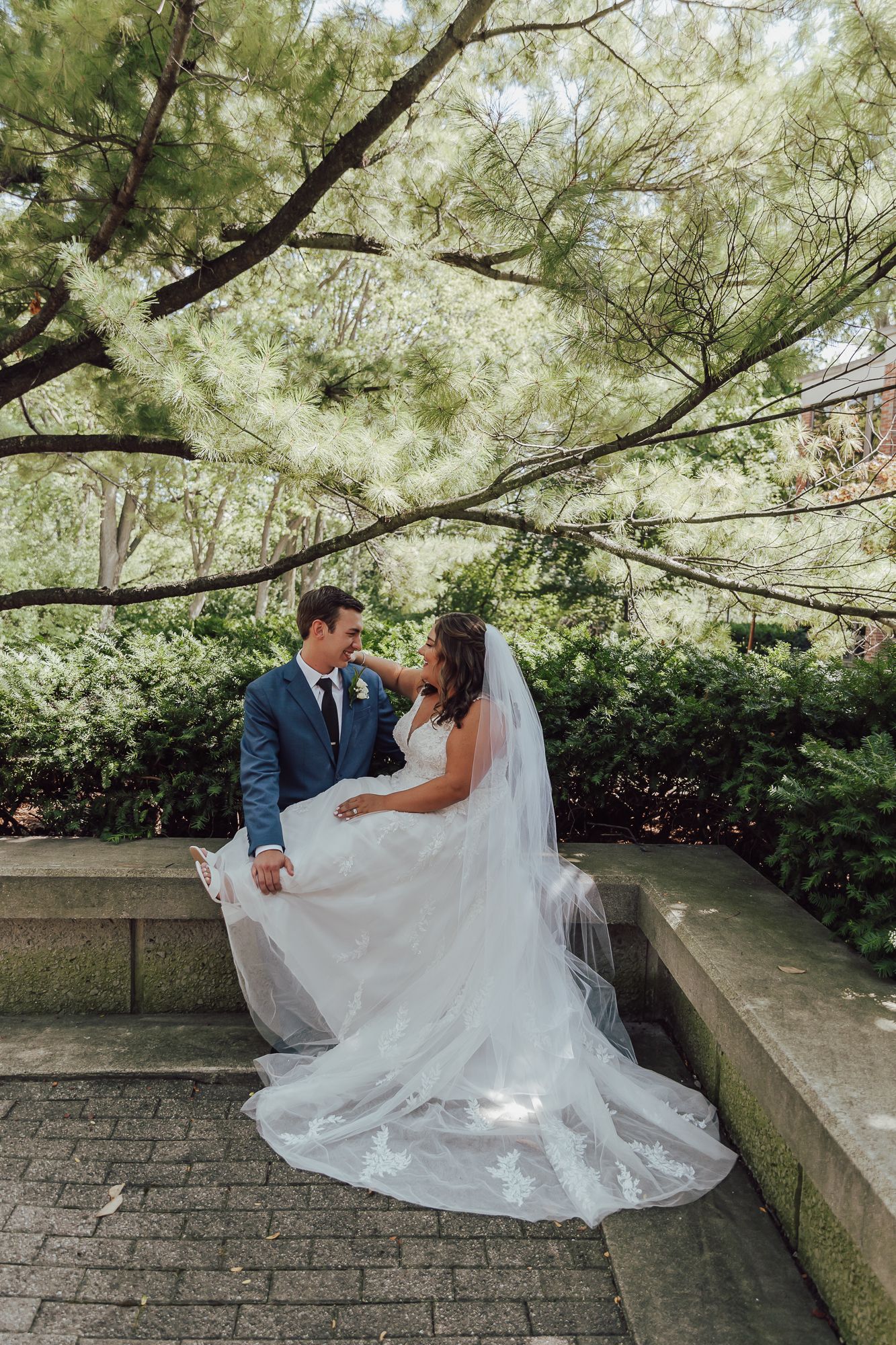 Newlyweds share an intimate moment under trees with bride's flowing white dress trailing on stone path.