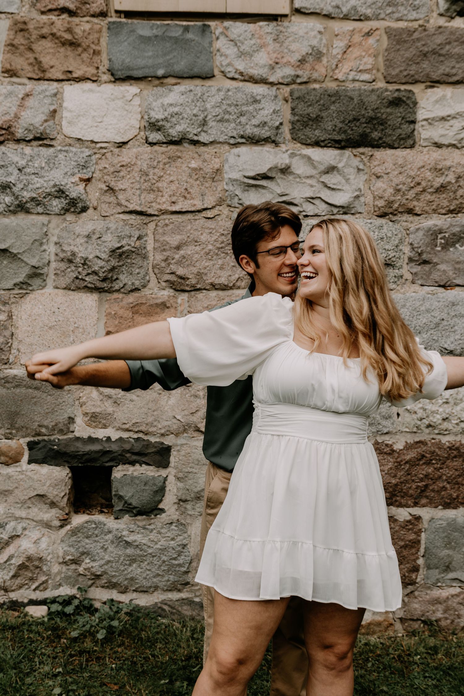 Couple in white dress and green shirt posing playfully against a stone wall in outdoor engagement photos.