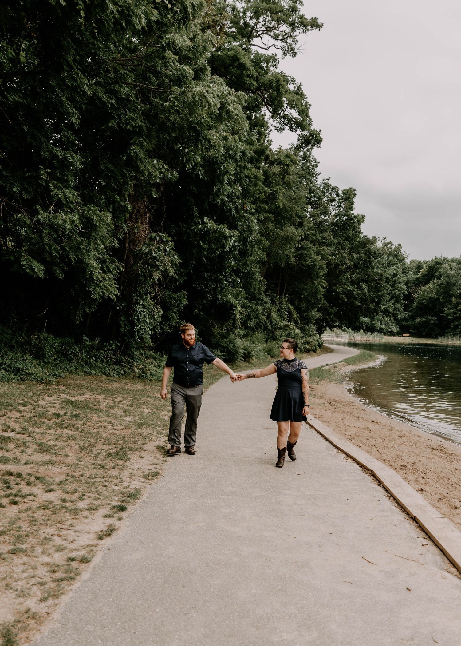 A couple holds hands while walking along a lakeside path on a cloudy day.