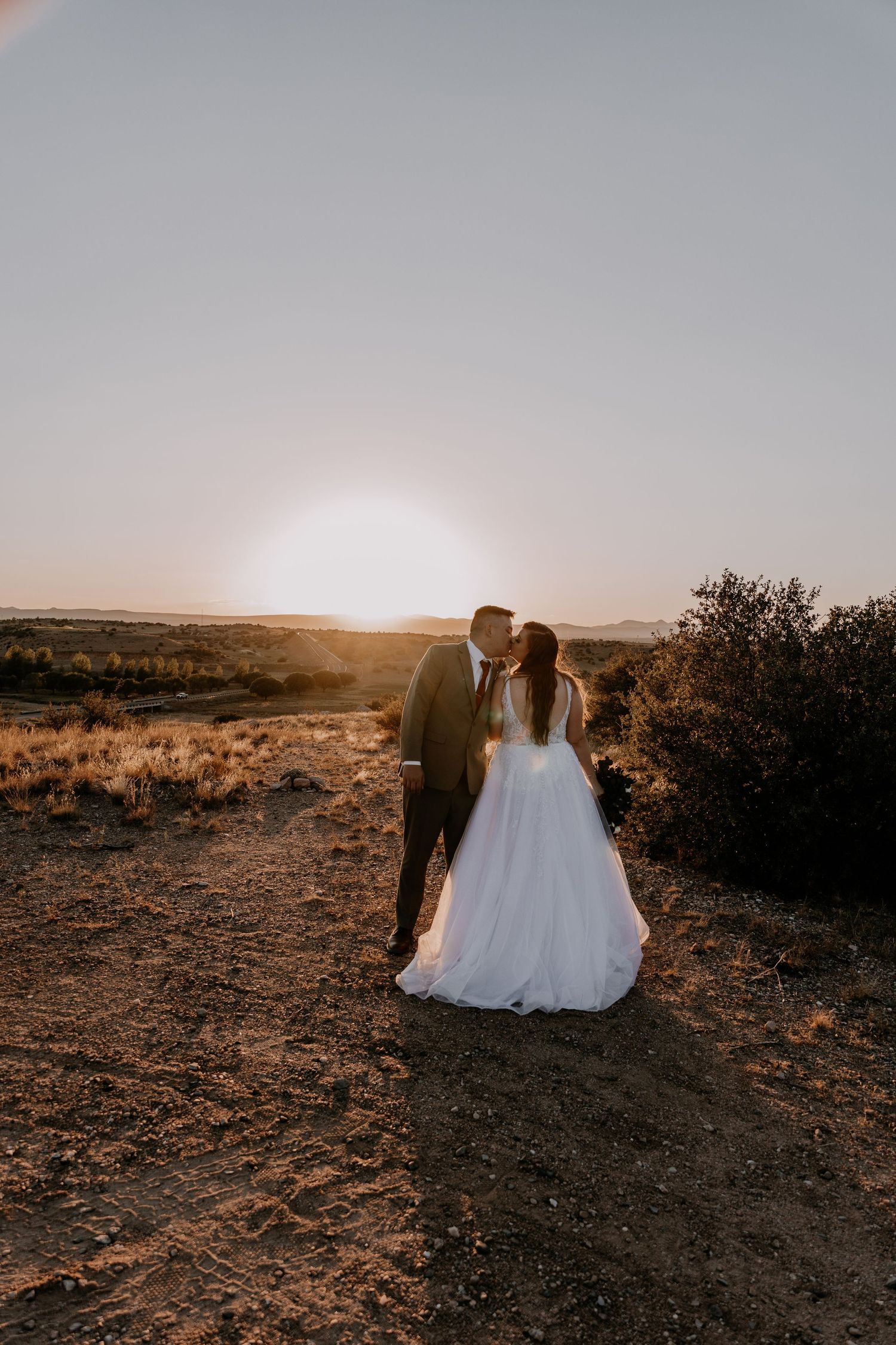 Couple in wedding attire walks hand in hand through rugged desert terrain at sunset.