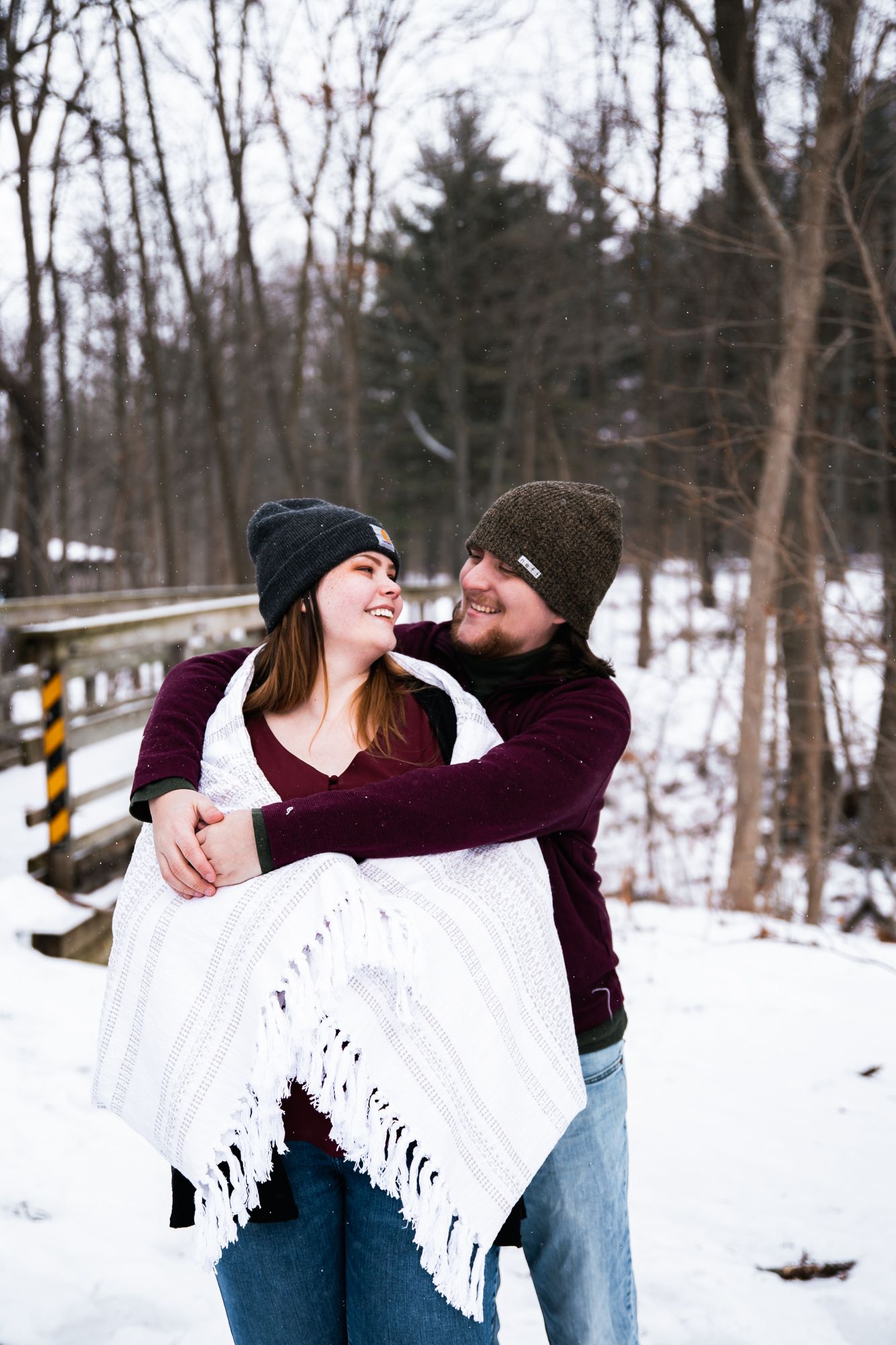 Couple wrapped in a white blanket sharing joyful moments in a winter forest setting.