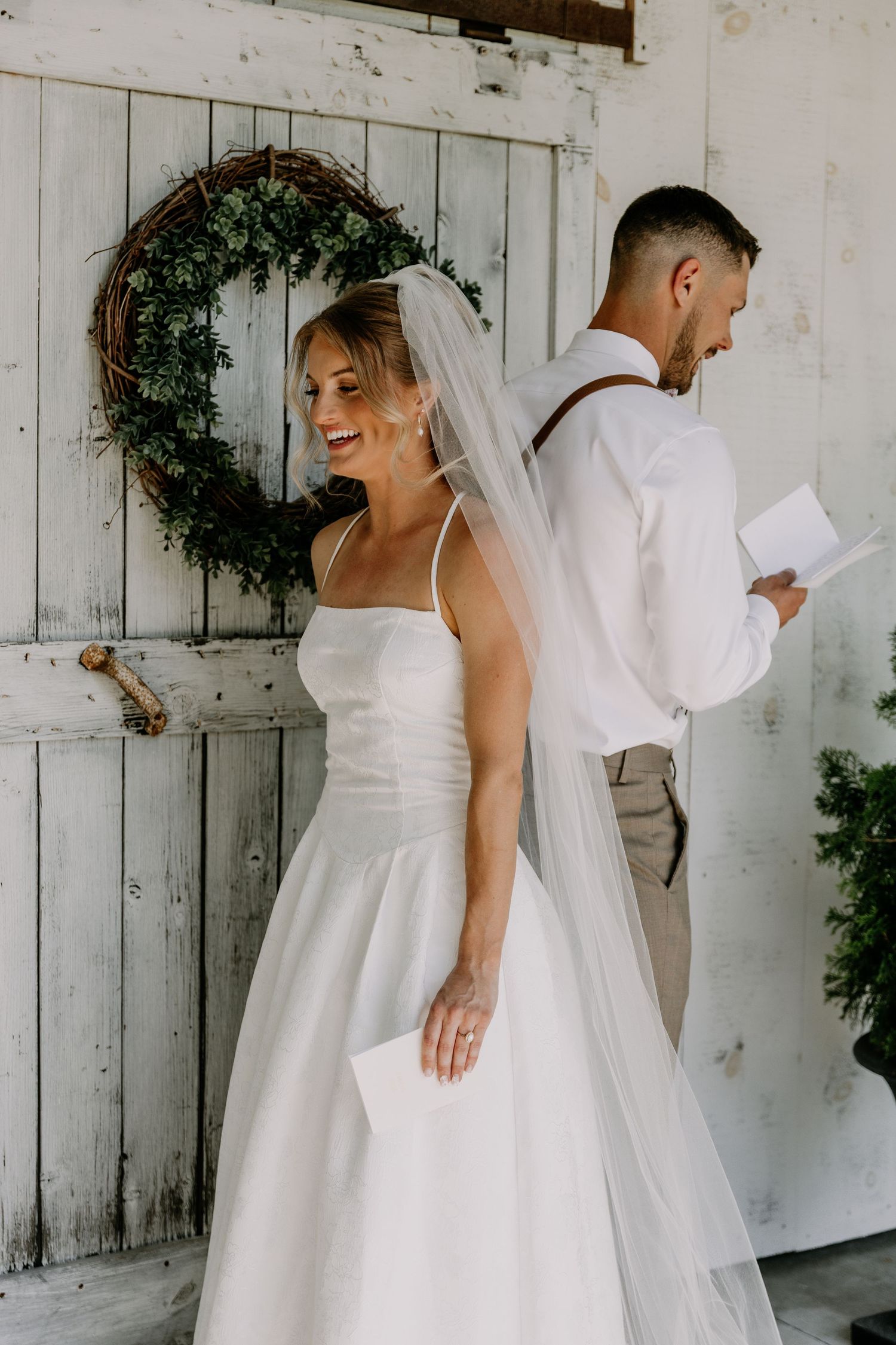 Bride in white gown and veil smiles while groom reads vows, standing back-to-back before rustic door with wreath.