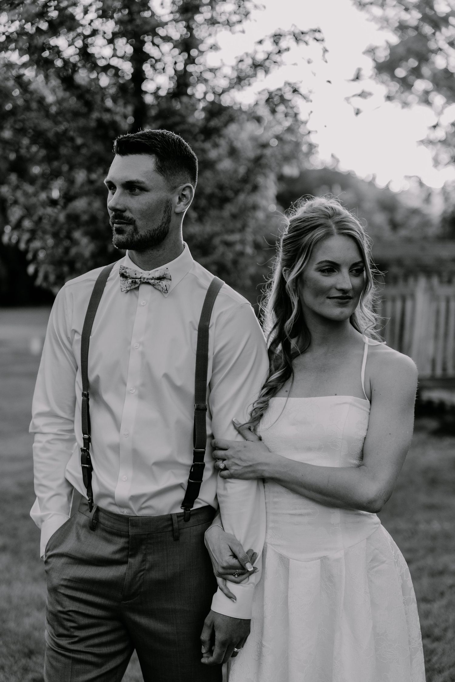 Black and white wedding photo of bride in white dress and groom in suspenders and bow tie posing outdoors.