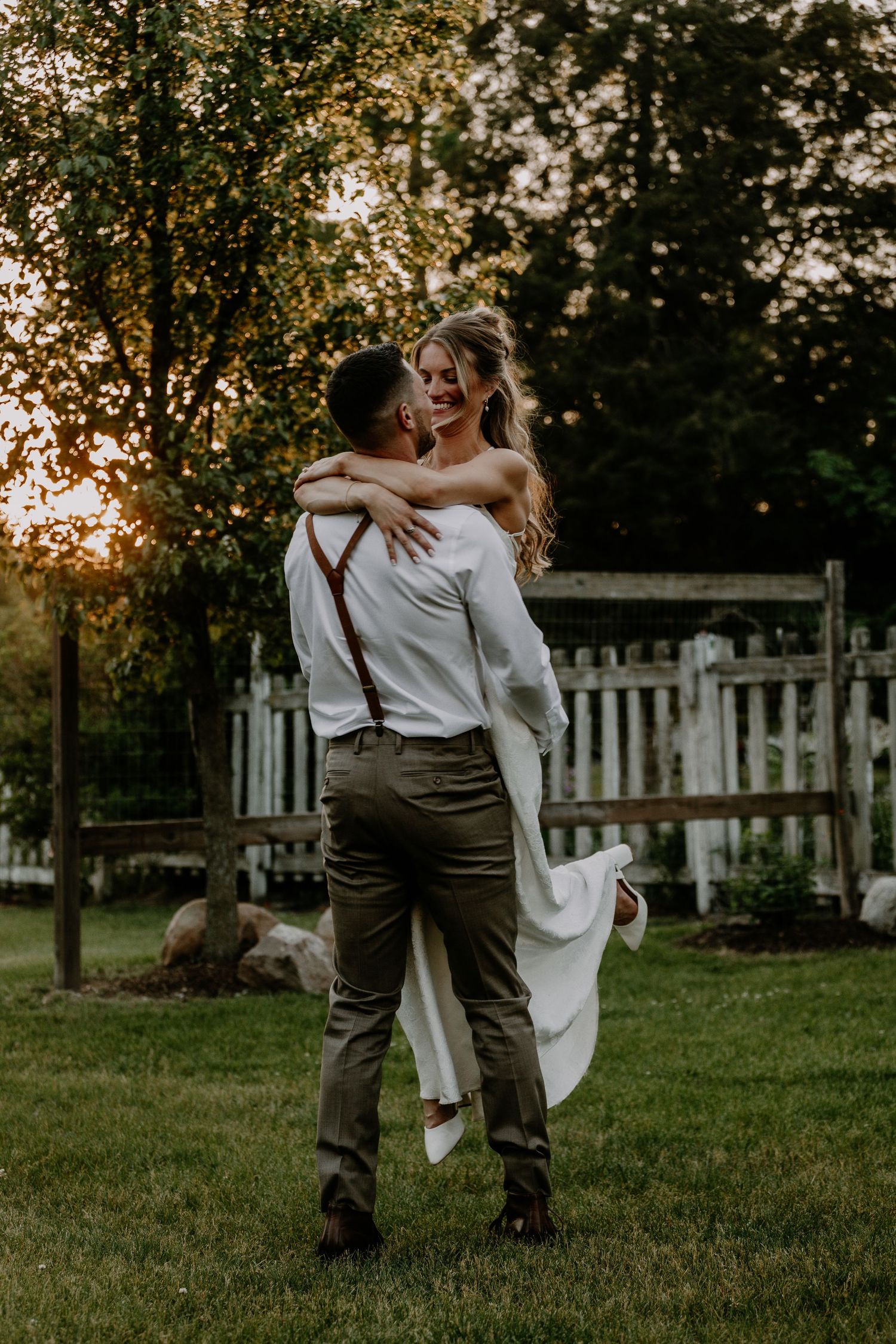 Groom lifts bride in white wedding dress at golden hour in a lush garden with a wooden fence backdrop.