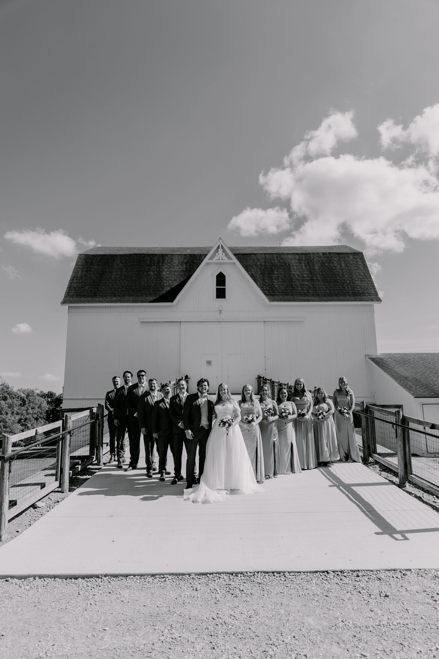 Wedding party poses in front of a white barn chapel on a sunny day.