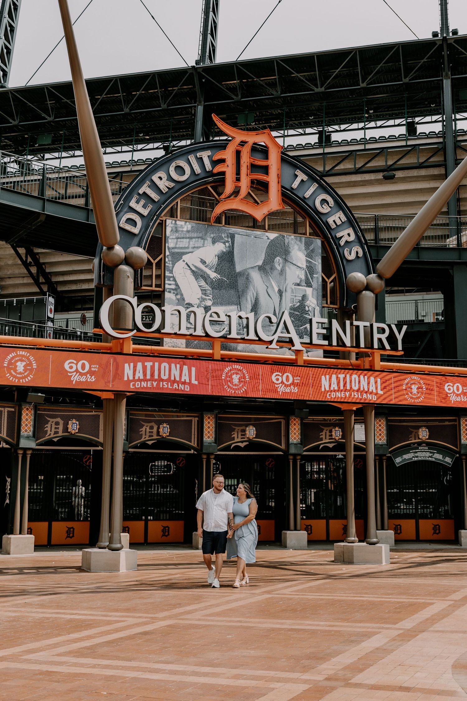 Two people walk outside Comerica Park baseball stadium entrance in Detroit featuring the iconic Tigers logo.
