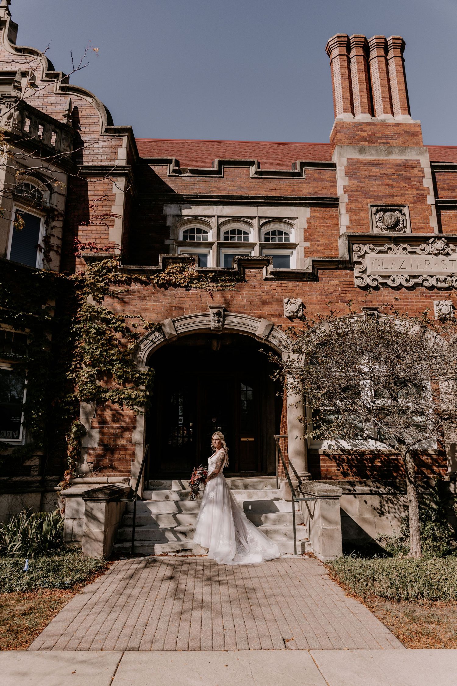 A bride in a flowing white gown stands in front of an elegant brick mansion entrance.
