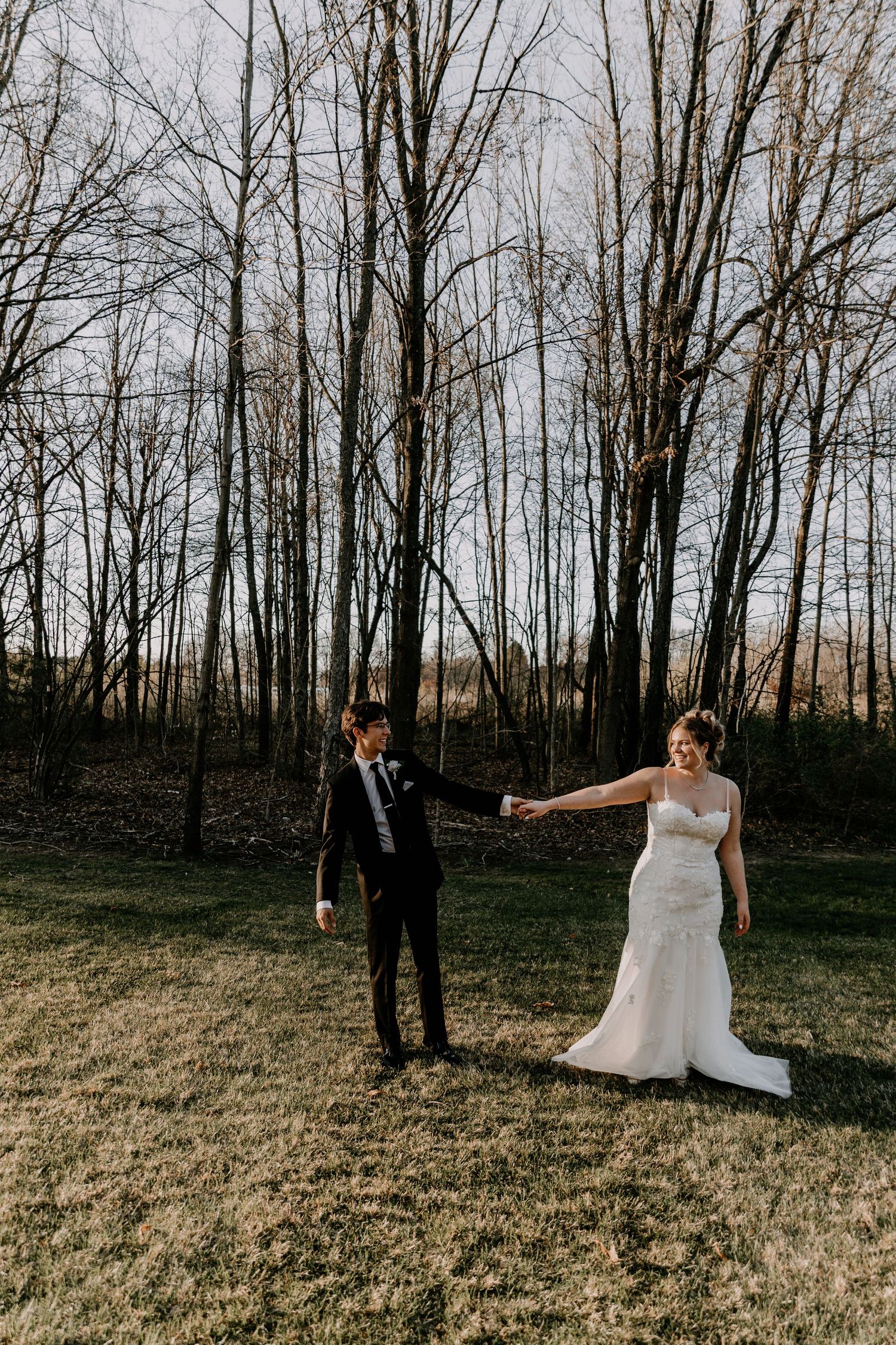 Newlyweds share playful moment in field with bare trees during golden hour.