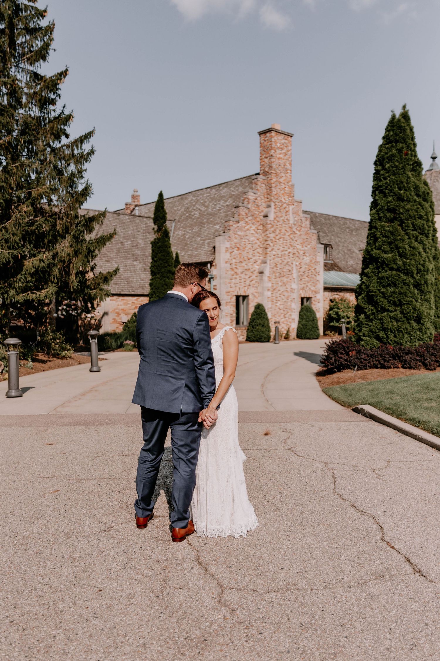 Newlyweds walking hand in hand on path outside stone church with tall cypress trees.
