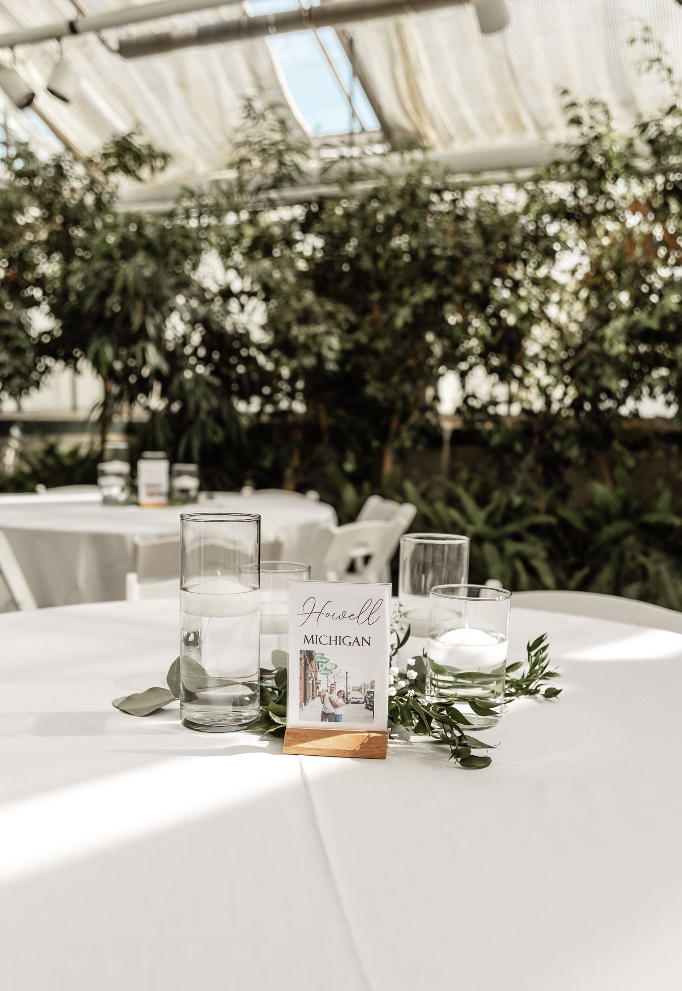 Elegant wedding reception table setting in greenhouse with white linens and simple centerpiece.