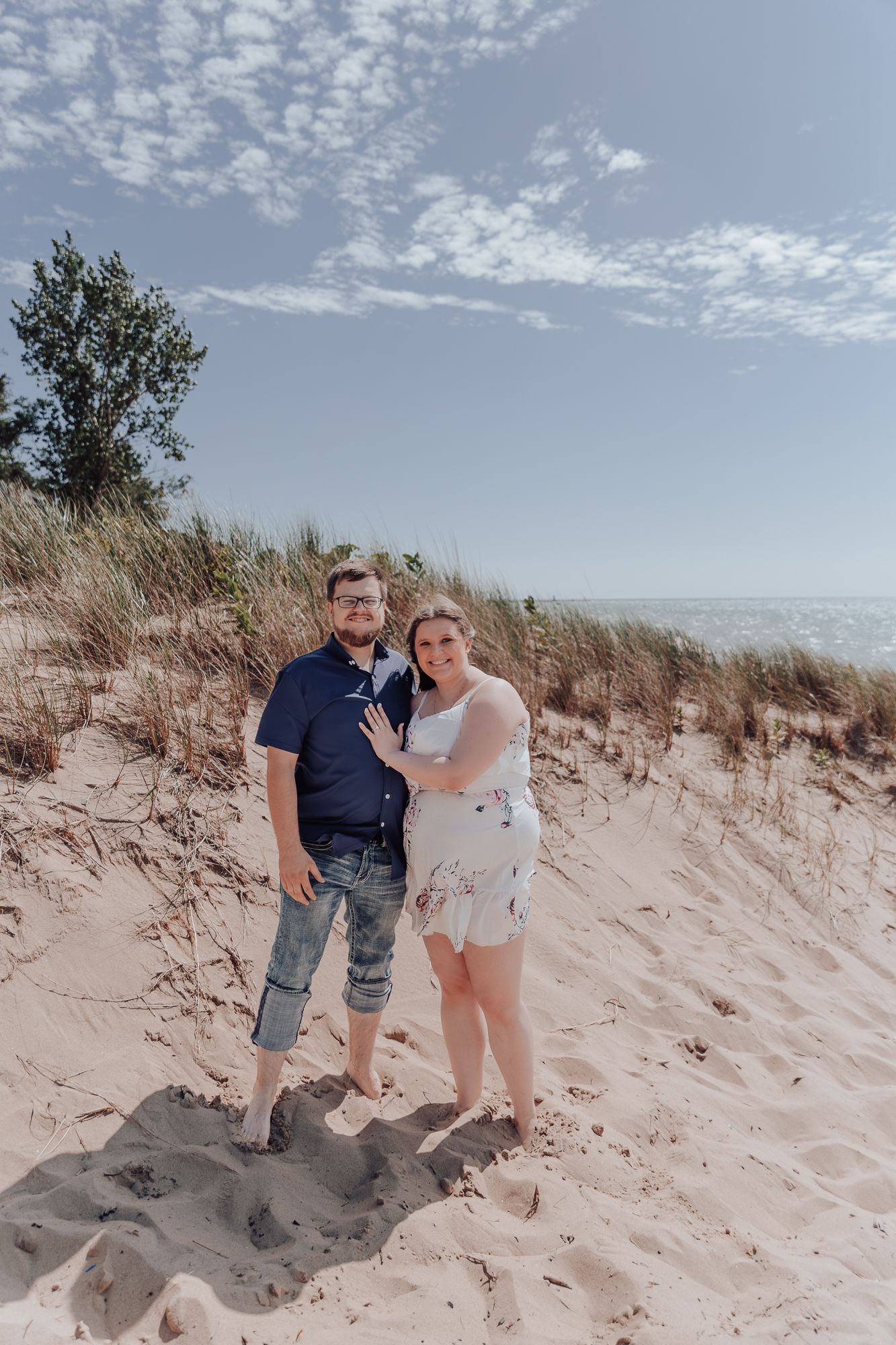 A couple poses together on sandy dunes with blue sky and coastal vegetation in the background.