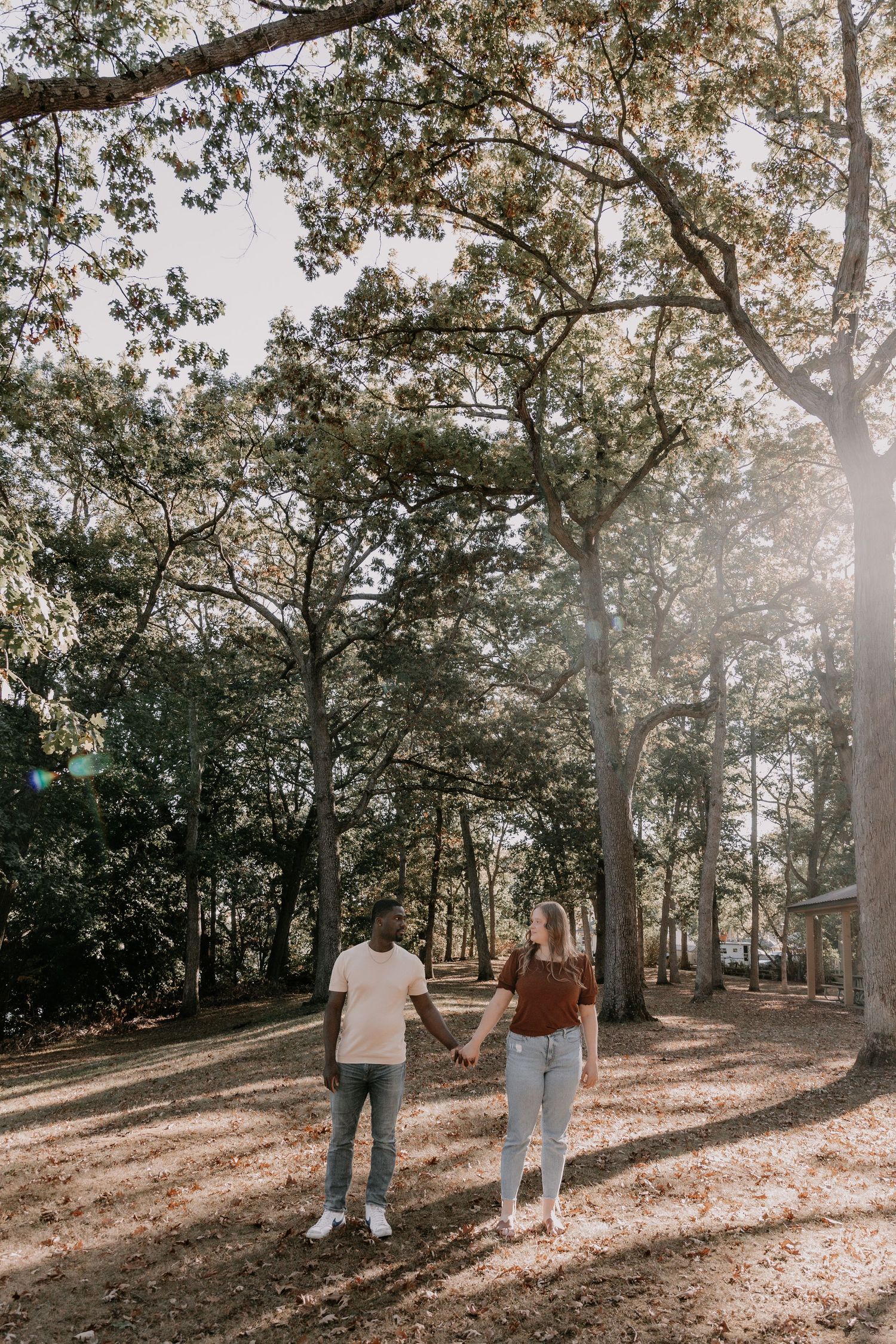 A couple walks through a sunlit wooded path with fallen autumn leaves creating a warm, romantic atmosphere.