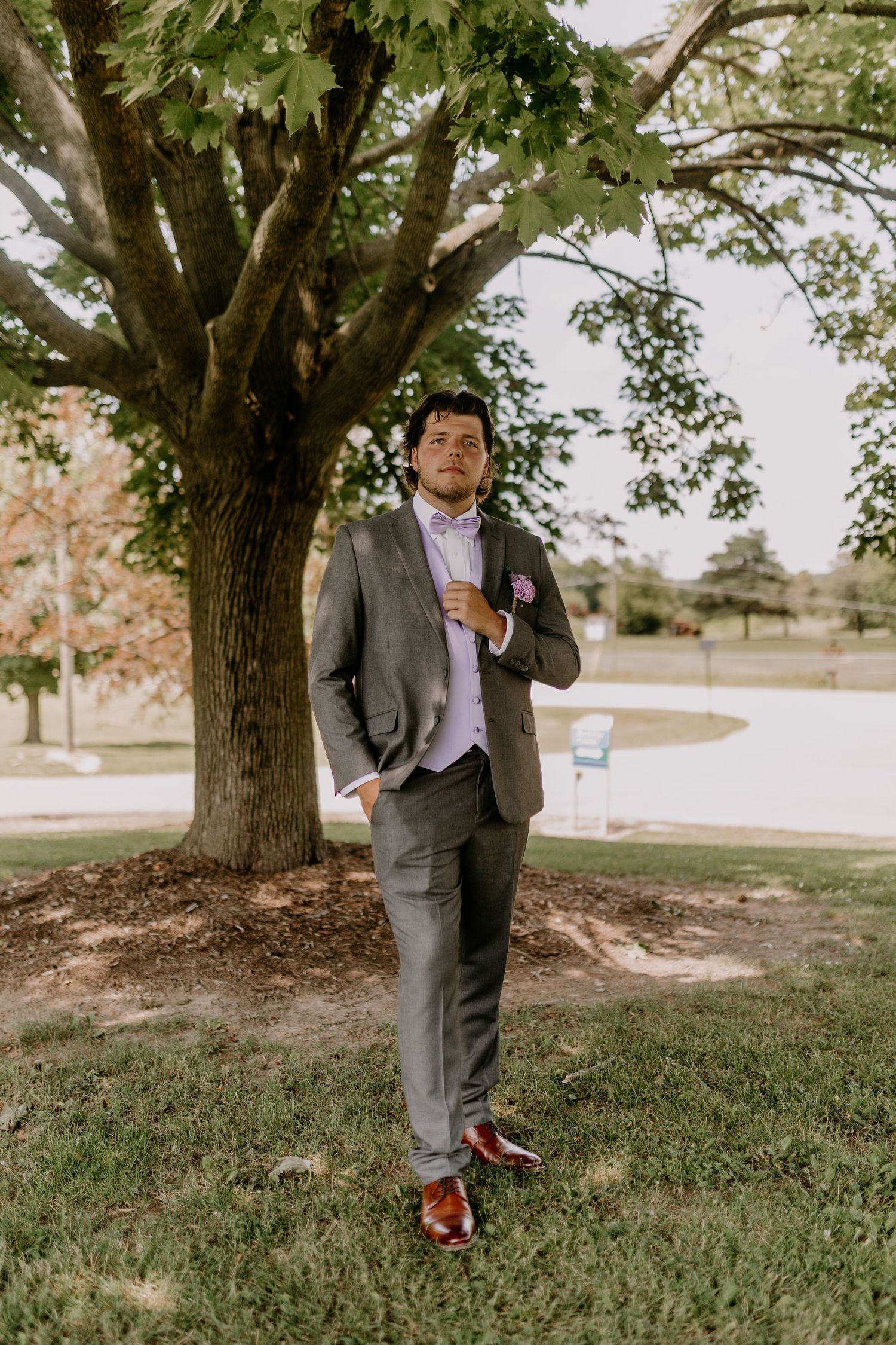 A dapper groom in a gray suit adjusts his tie while standing under a large tree outdoors on his wedding day.
