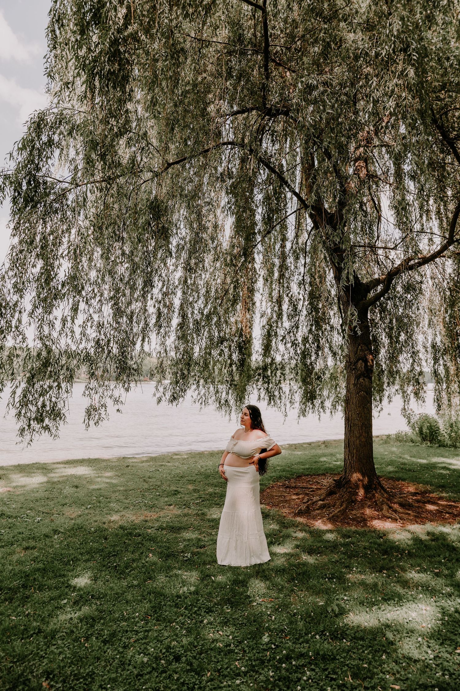 Pregnant woman in white off-shoulder gown standing under a weeping willow tree by a serene lakeside on a cloudy day.