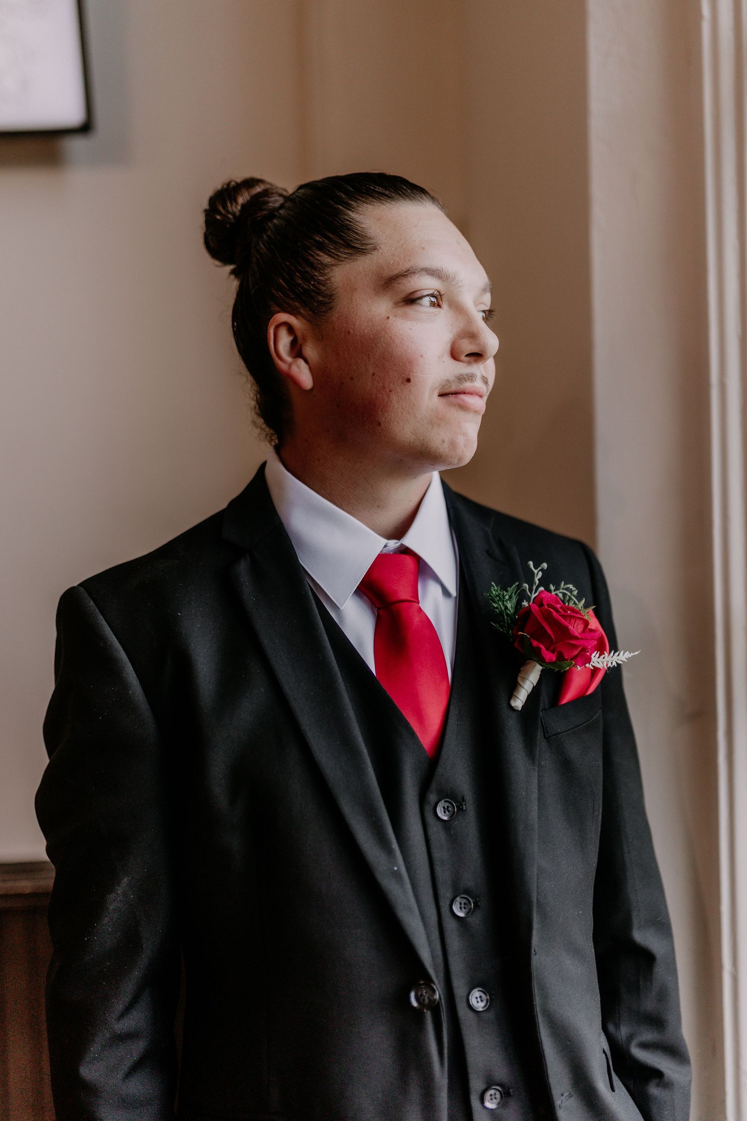 Young groom in black suit with red tie and rose boutonniere gazing thoughtfully during wedding preparations.