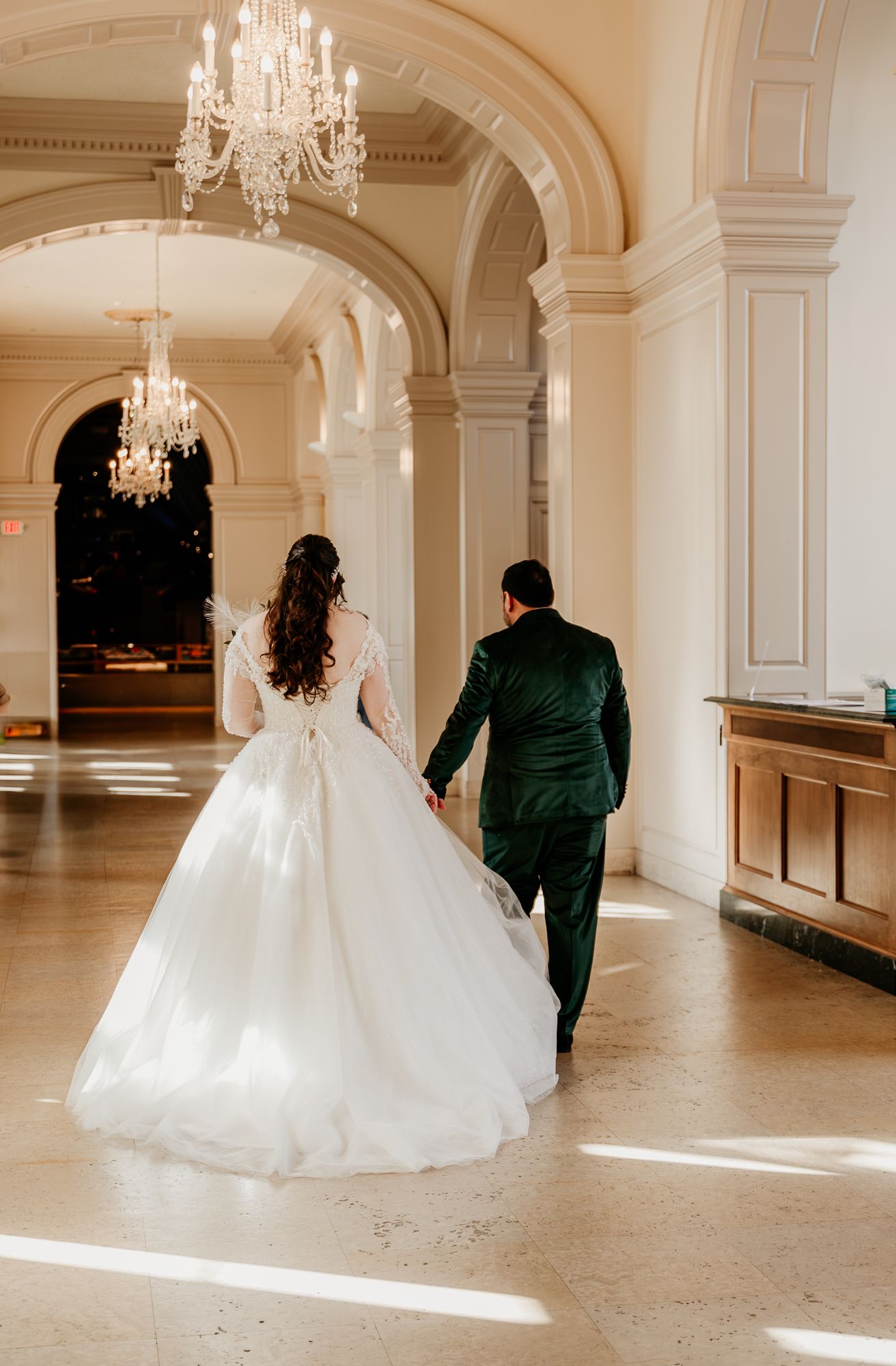 A elegant couple in wedding attire walking down a grand hallway with crystal chandeliers and arched architecture.