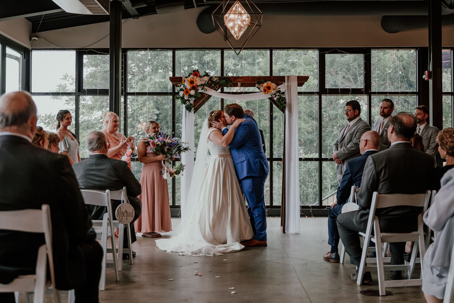 A wedding ceremony in a modern venue with large windows and geometric chandeliers as a couple exchanges vows.