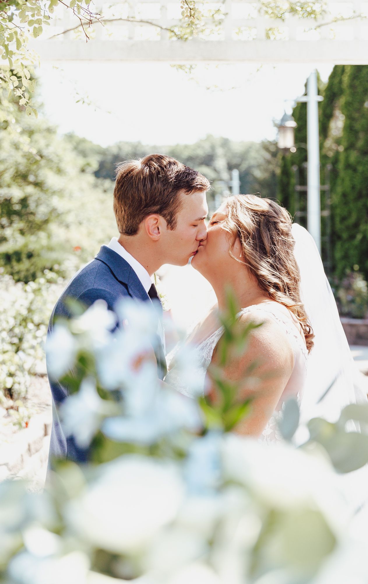 Romantic garden kiss between newlyweds framed by soft white flowers and greenery.