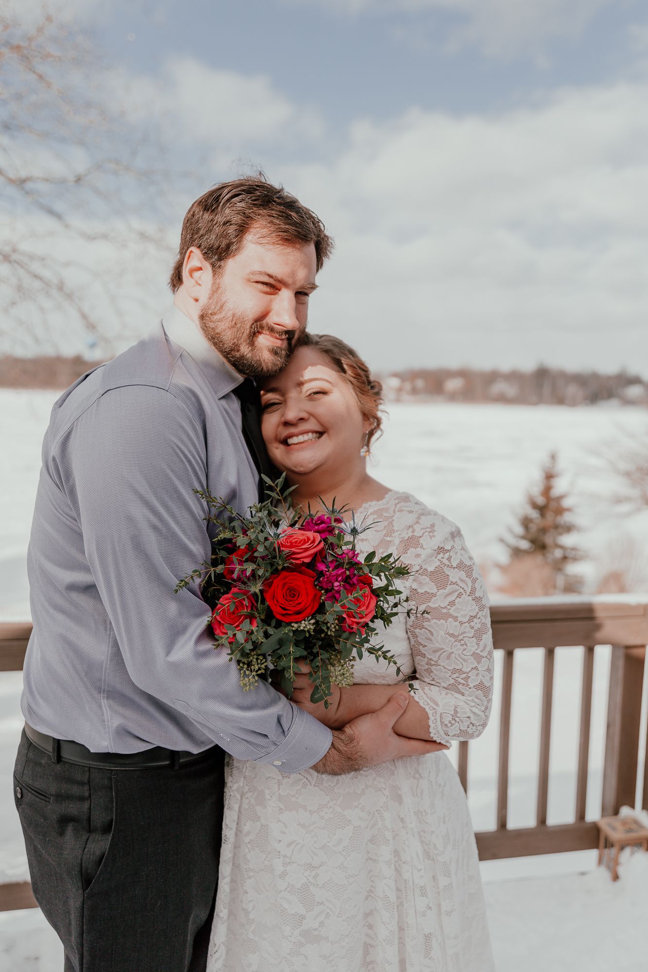 A winter wedding scene with a couple embracing outdoors holding a red rose bouquet.