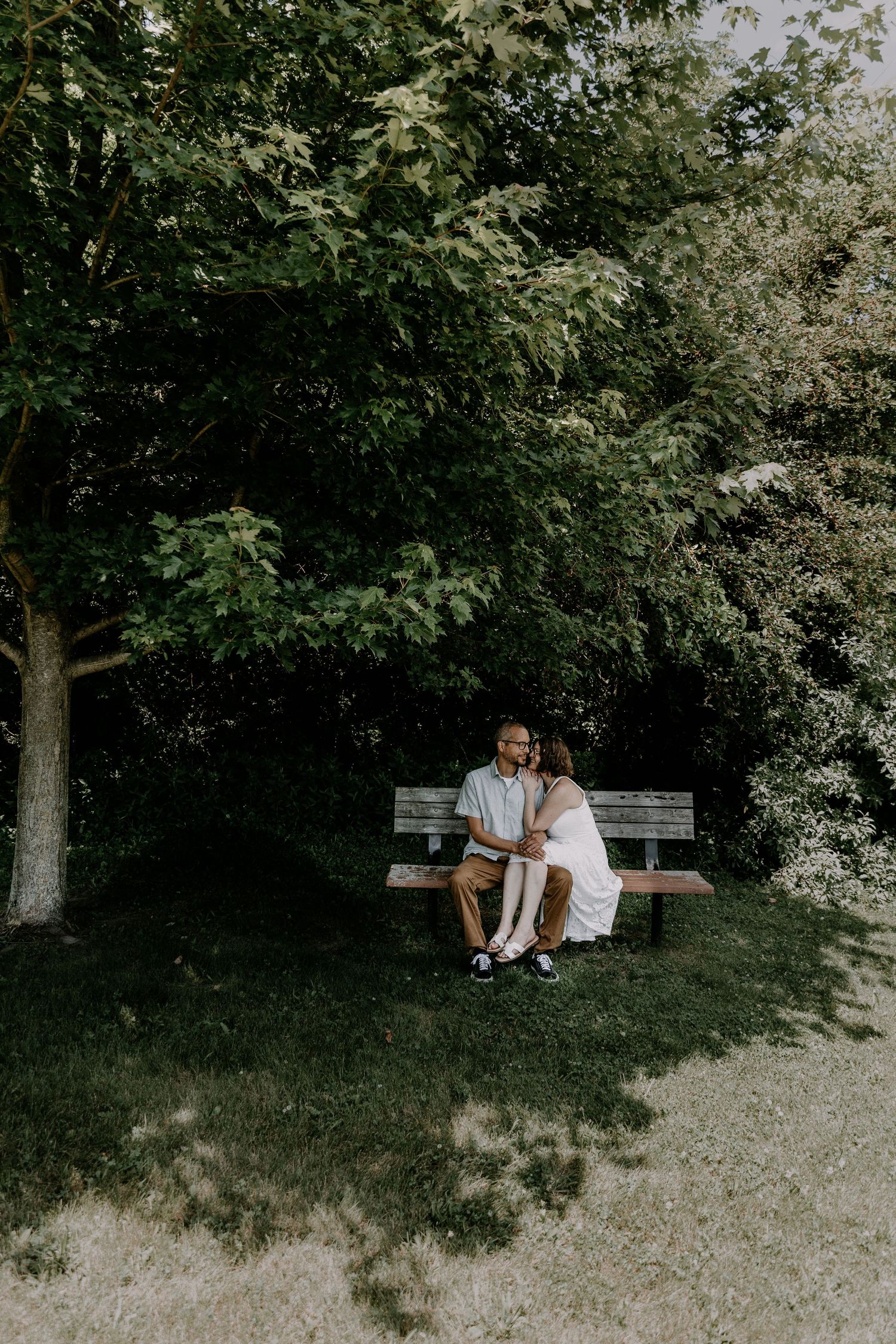 A couple sits together on a wooden bench in a serene garden setting surrounded by lush green trees and foliage.