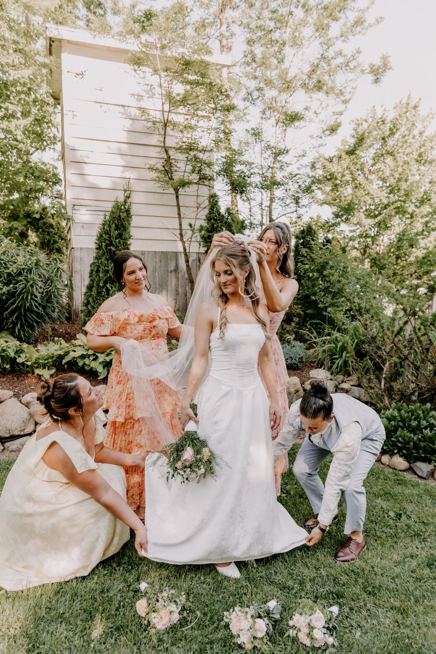 Bridesmaids in floral dresses help arrange bride's veil and train on green lawn during outdoor wedding preparation.