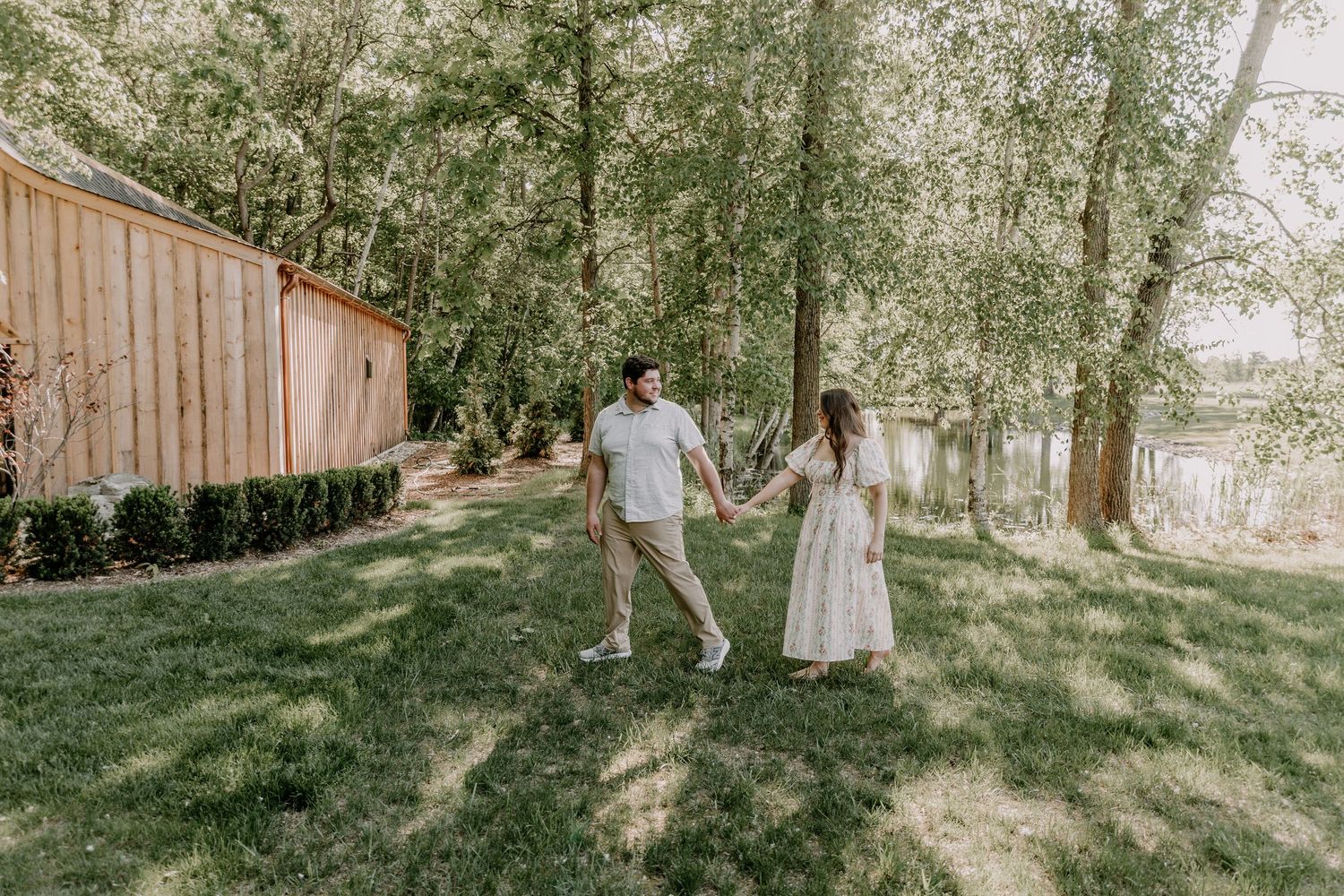 Couple holding hands and dancing in a lush green backyard near a wooden barn surrounded by tall trees.