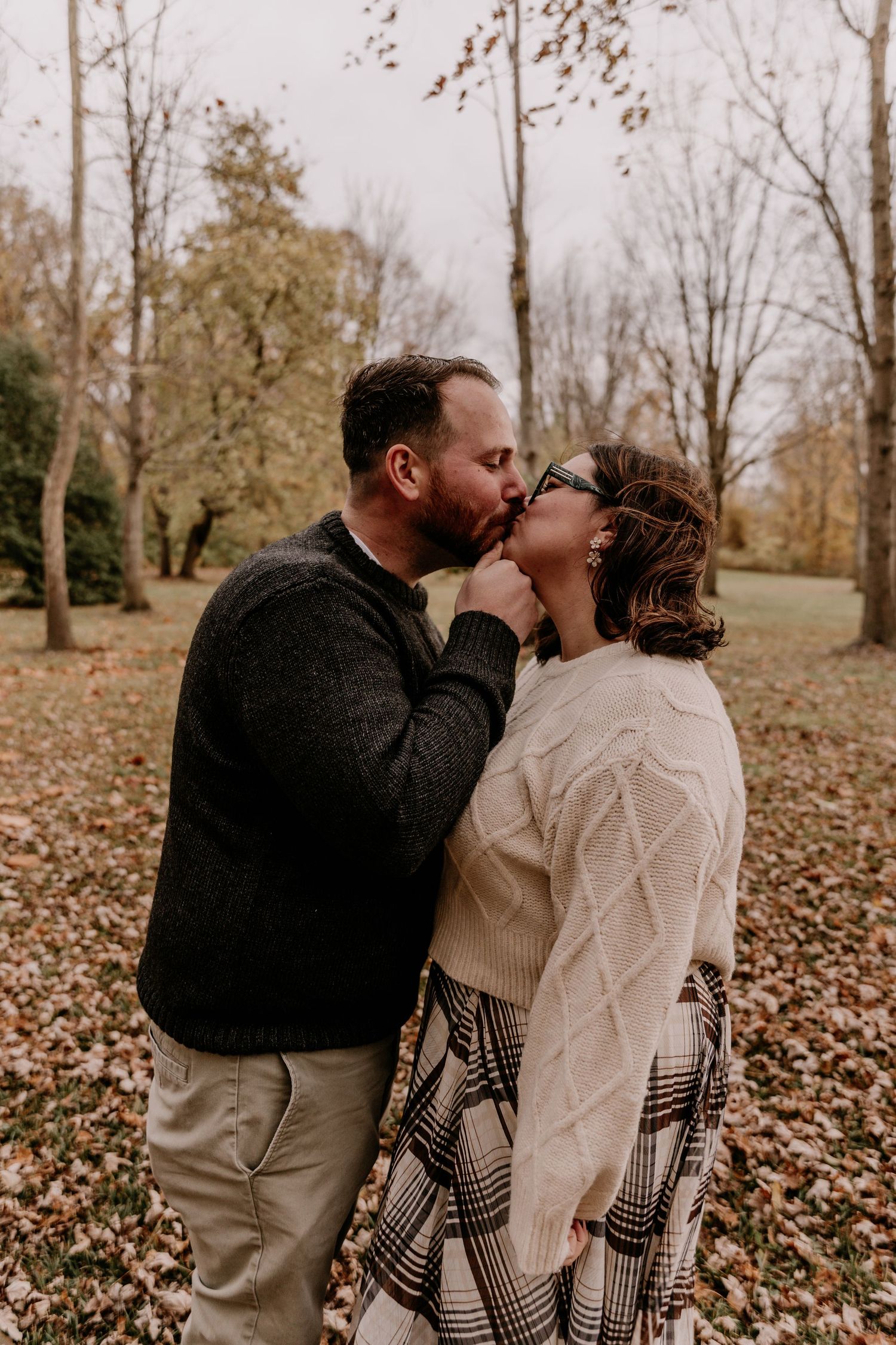 A couple shares romantic kisses in a fall setting with leaves scattered on the ground during a photo session.