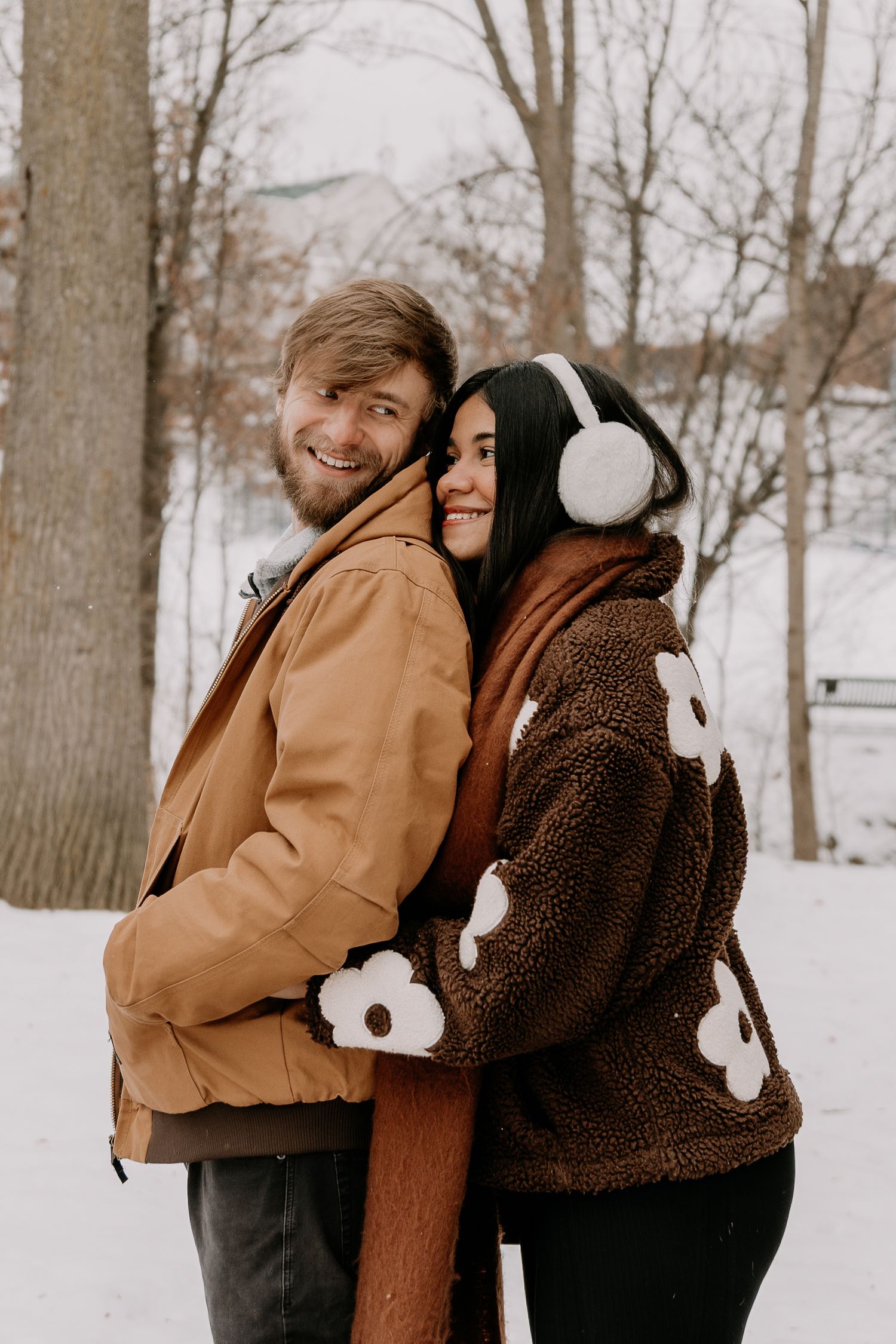 Two people in winter coats embrace and laugh together in a snowy landscape with bare trees in the background.
