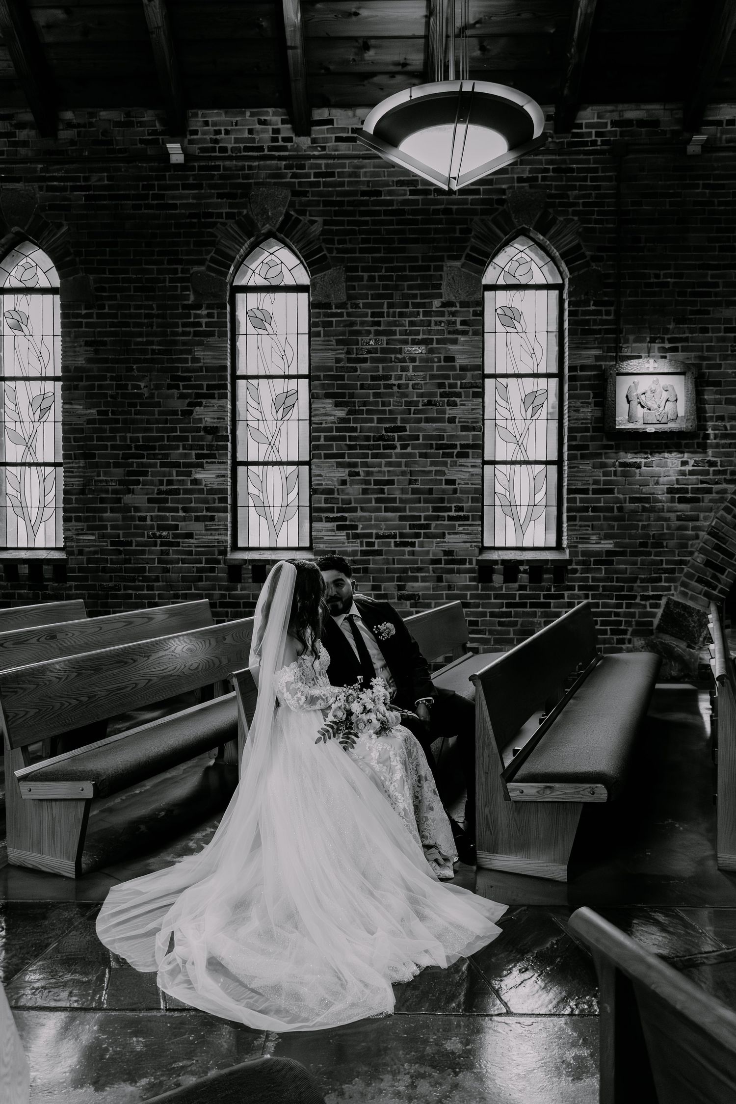 Black and white photograph showing a romantic wedding moment inside a historic brick church with arched windows and wooden pews.