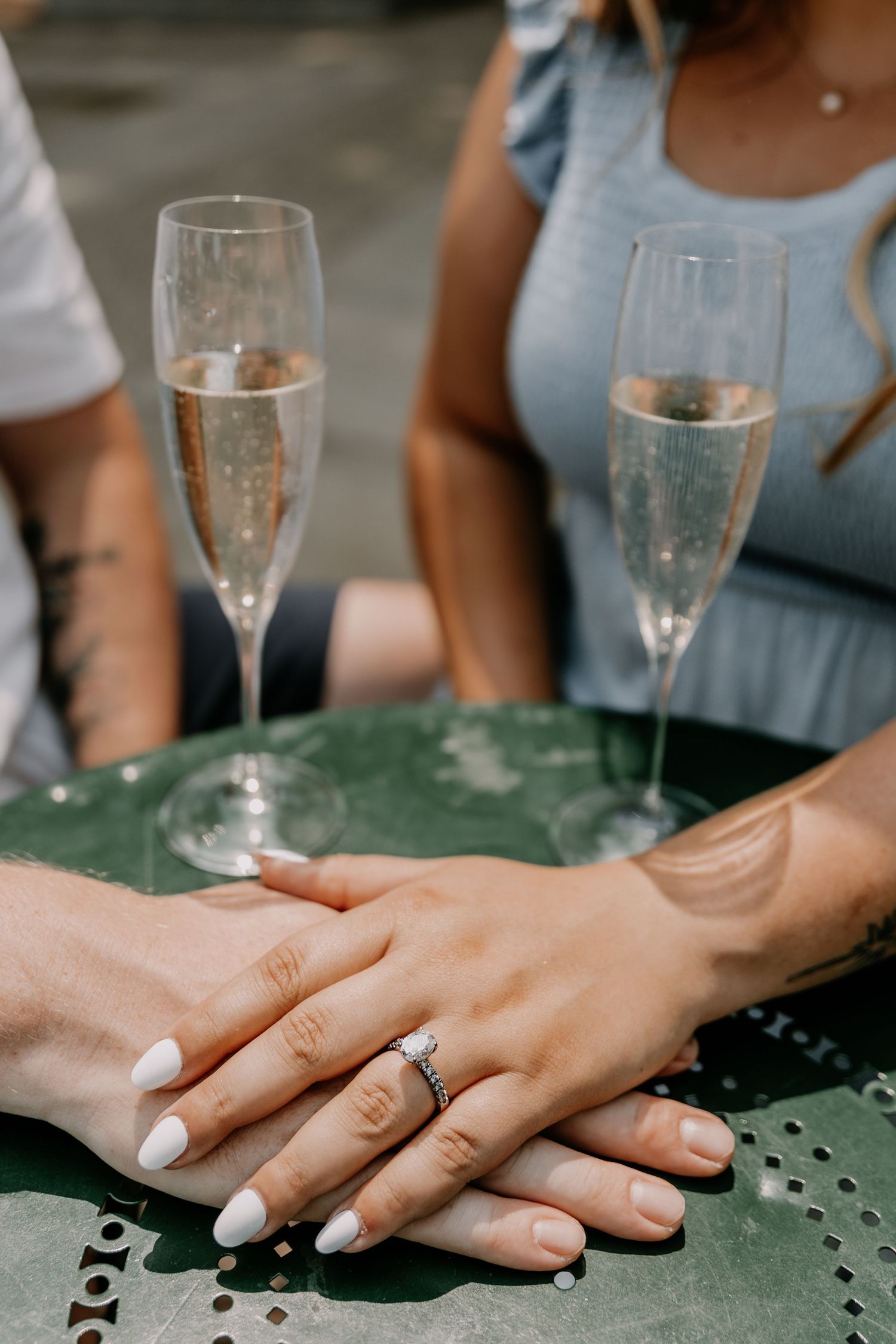 Two hands with white nail polish and engagement ring rest on green table beside champagne flutes during celebration.