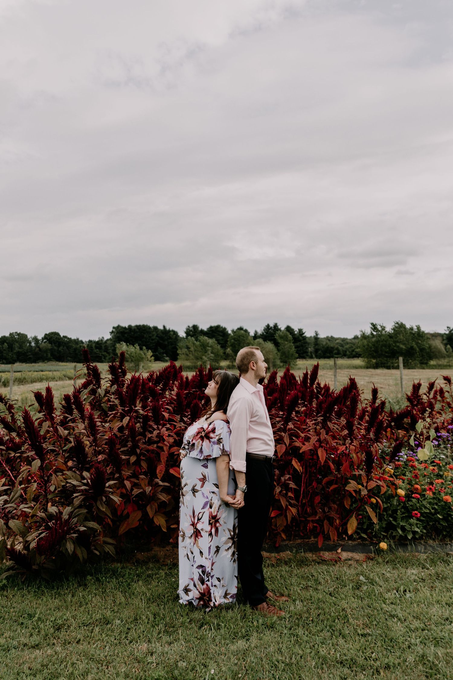 Two women in floral dresses standing near a row of vibrant red plants in a garden on an overcast day.