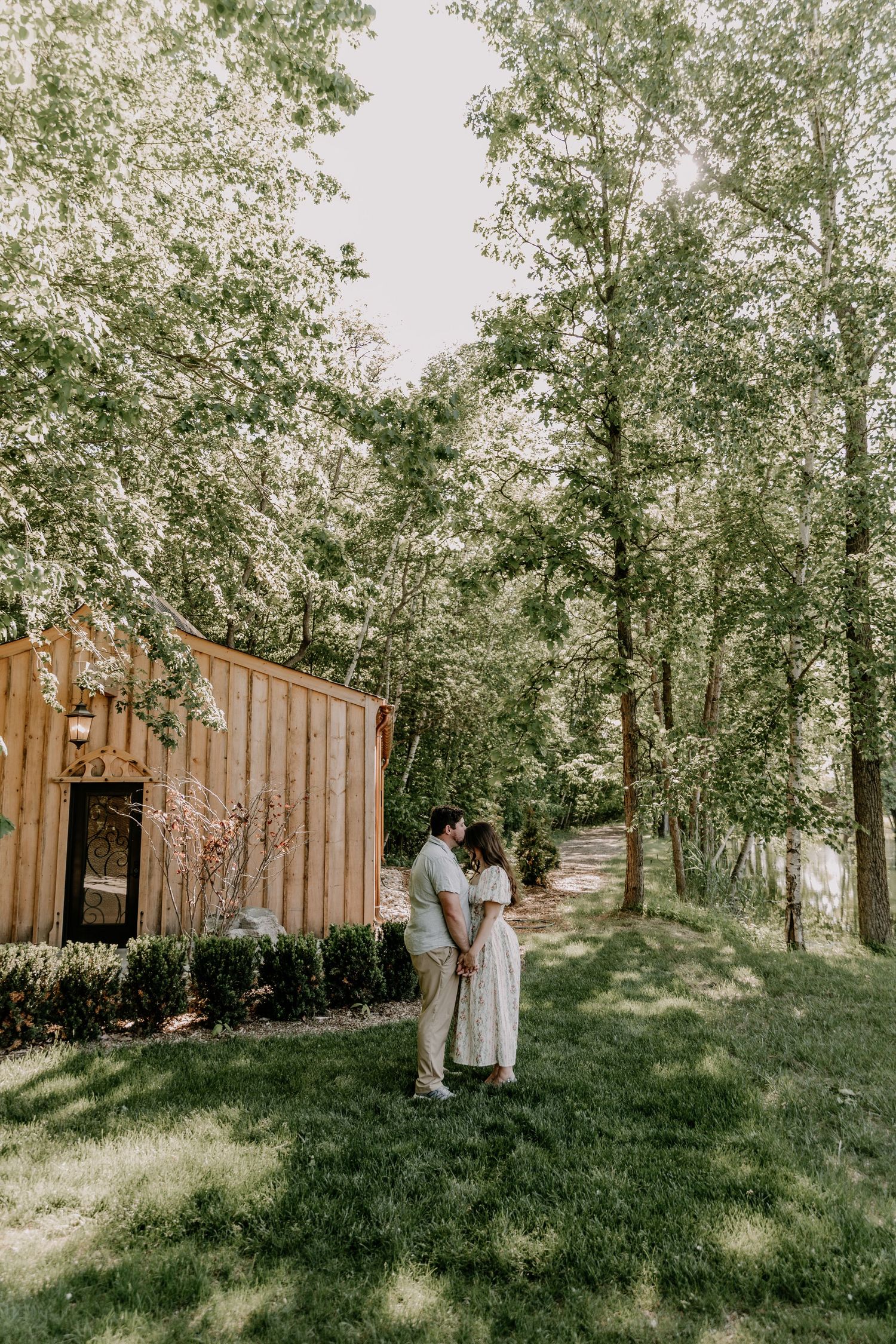 Couple sharing a tender moment on a lush green lawn beside a wooden cabin surrounded by tall trees.