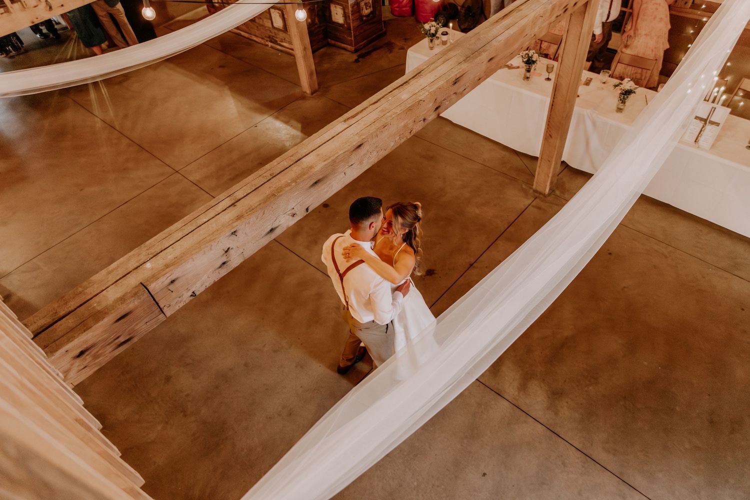 Aerial view of a bride and groom sharing a romantic moment on a rustic barn dance floor with white fabric draping.