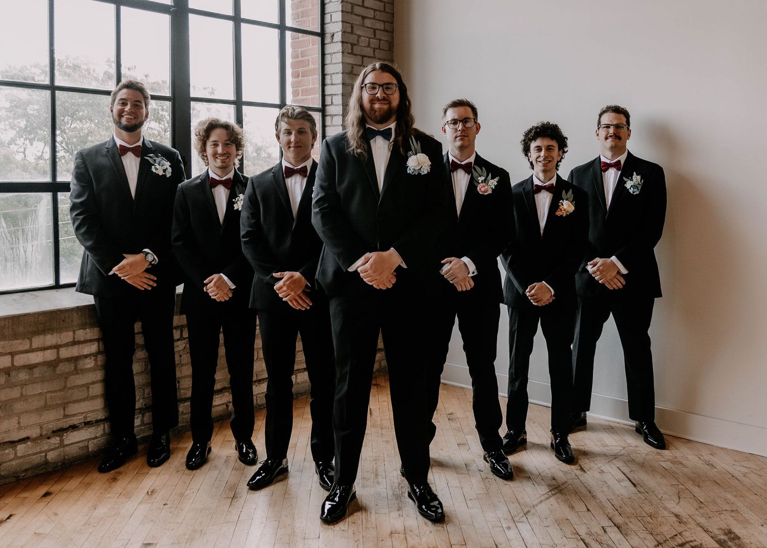 Wedding party members in formal attire stand in formation by large windows.
