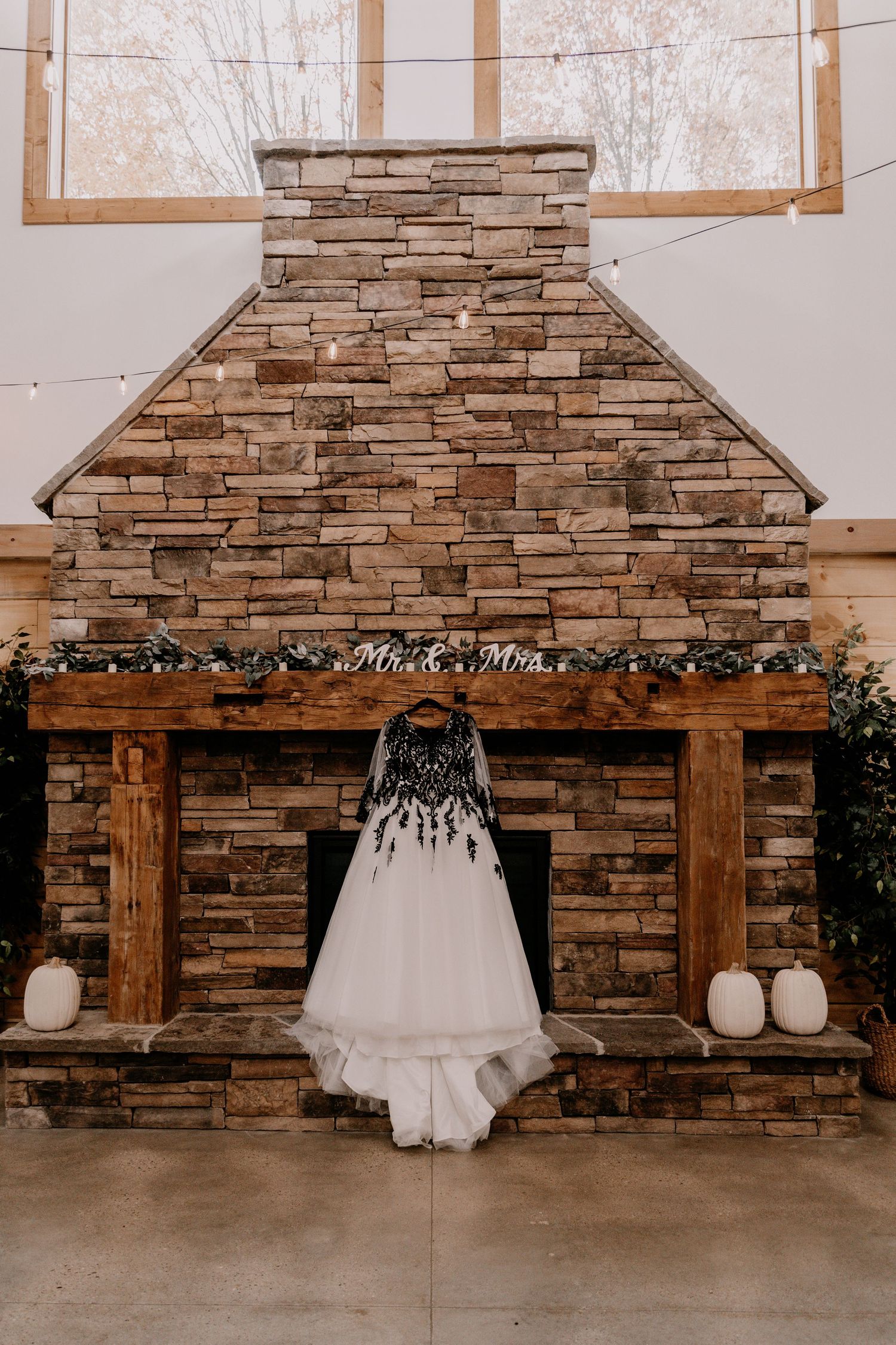 A white wedding dress hangs on a stone fireplace decorated with greenery and white pumpkins.