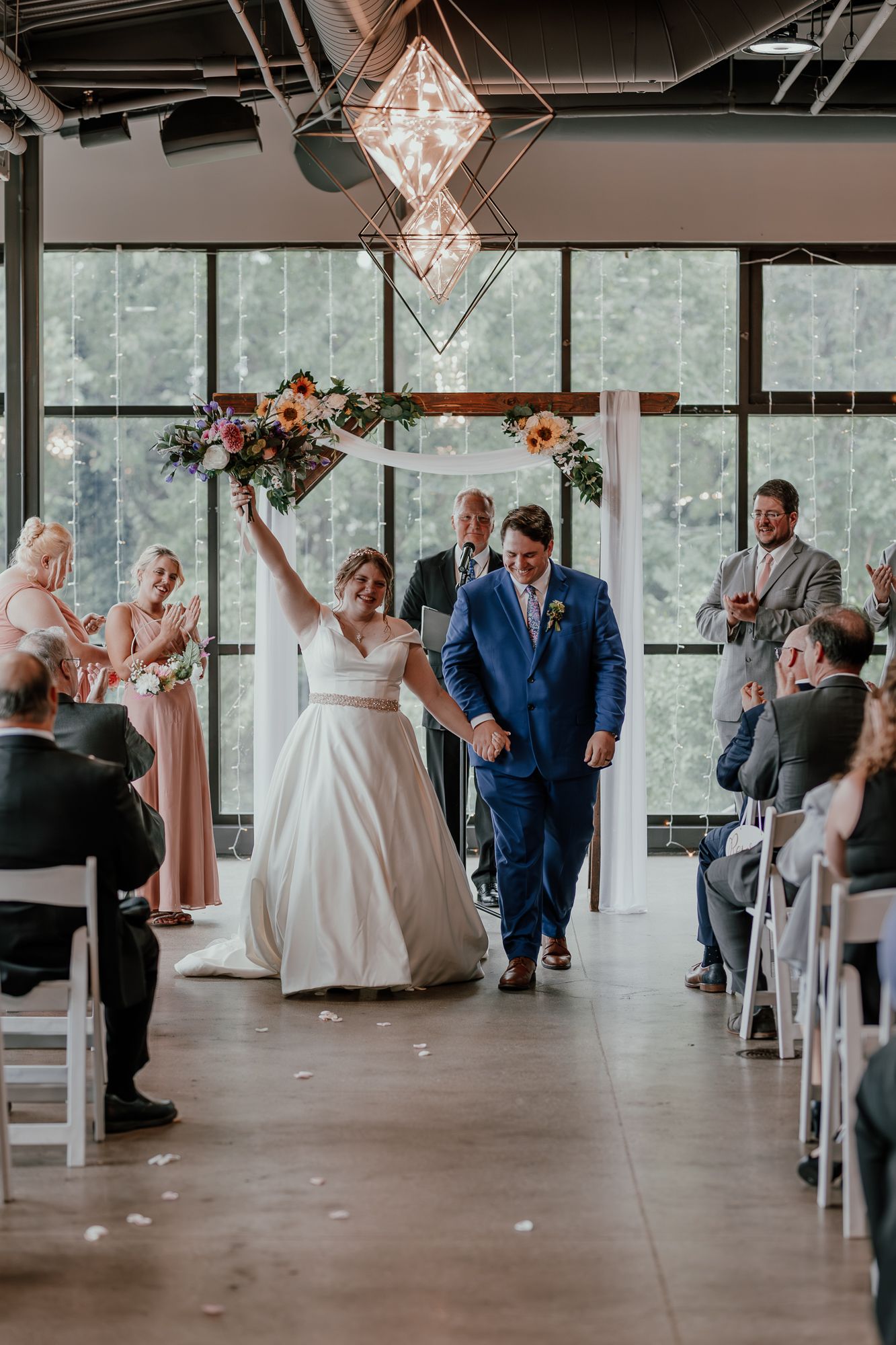 A joyful wedding ceremony moment under a floral arch with geometric lighting in an industrial space.