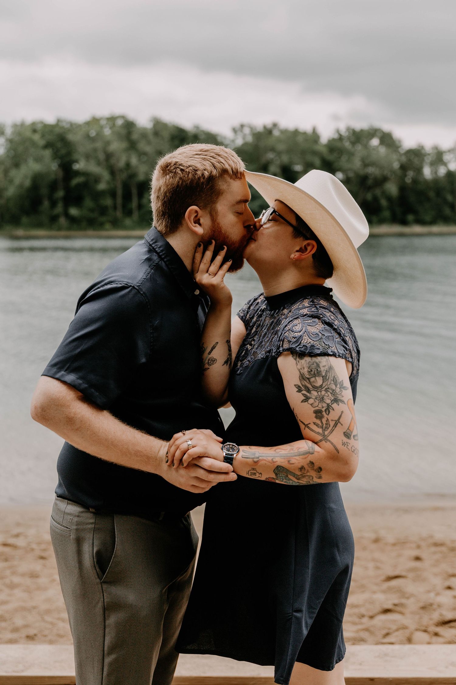 A couple shares a tender moment along a lakeside beach under cloudy skies.