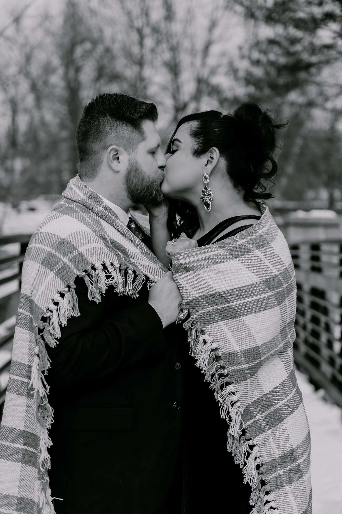 A couple sharing an intimate moment wrapped in a plaid blanket on a winter bridge.