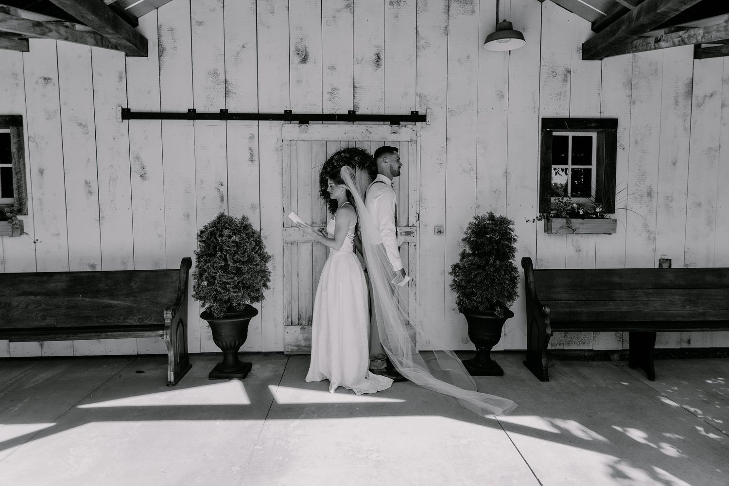 Black and white photo of a bride and groom standing back-to-back before a rustic barn door on their wedding day.