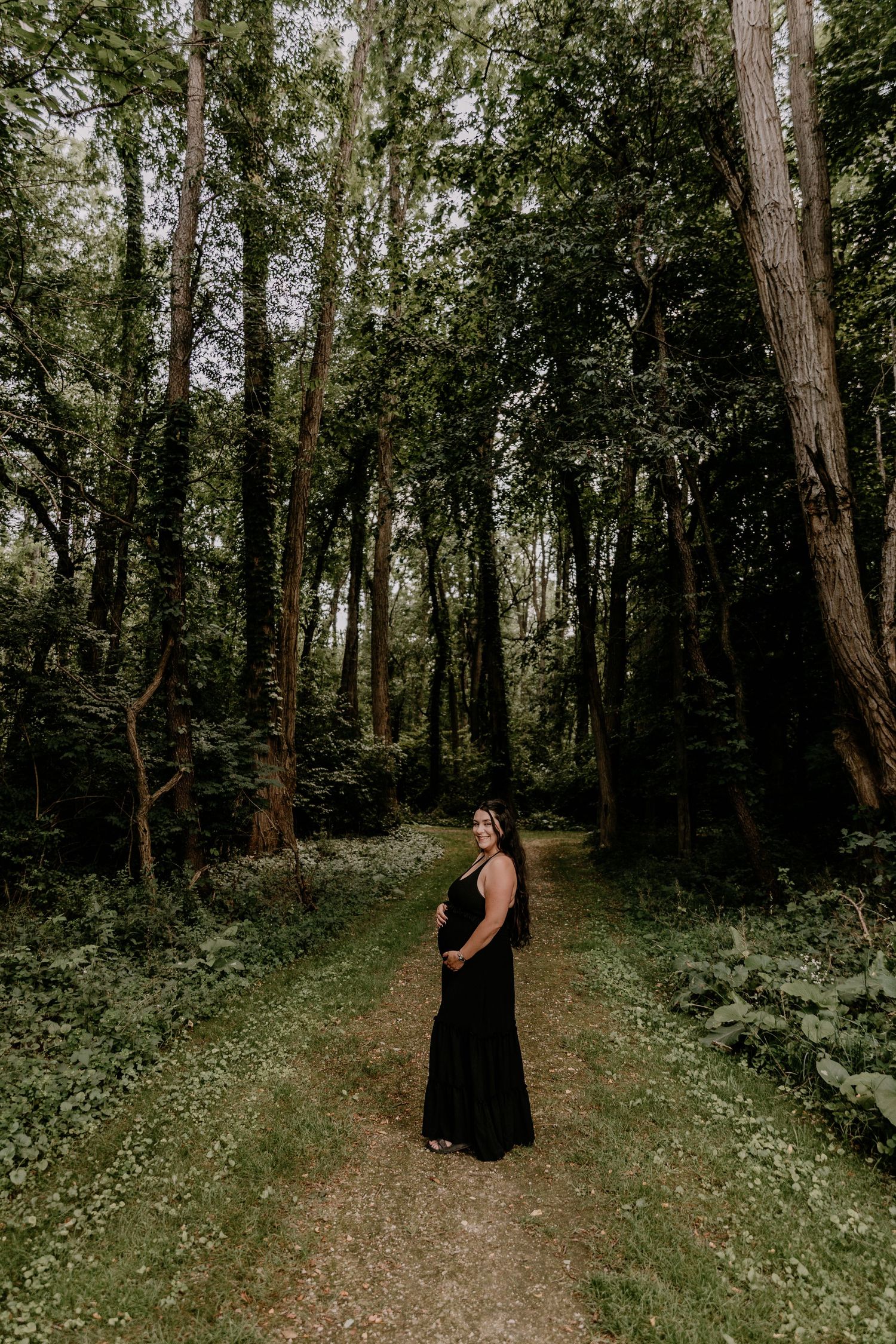 Pregnant woman in black maxi dress standing on forest path surrounded by tall trees and lush green foliage.