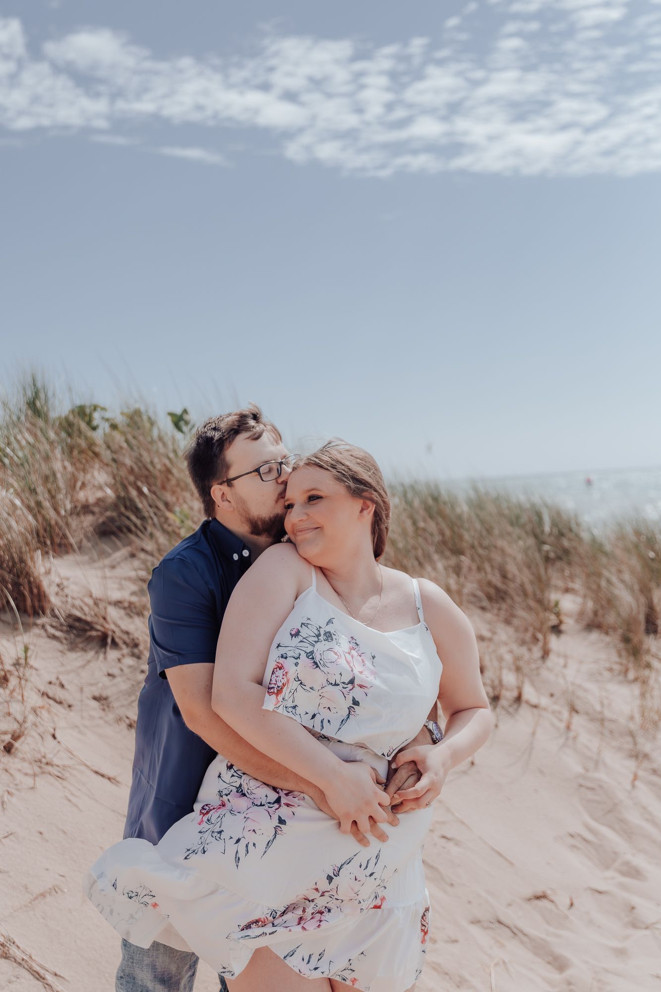 A romantic embrace captured on sand dunes with wispy clouds in the summer sky.