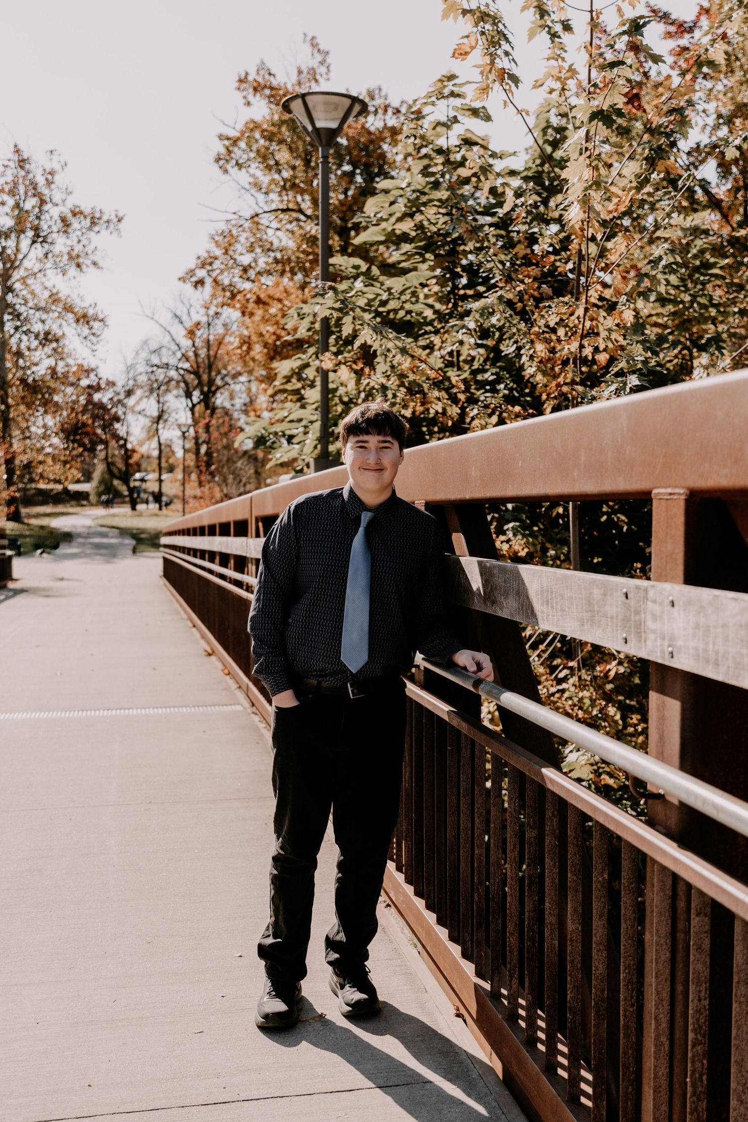 Person in formal black attire standing on wooden bridge walkway during autumn season.