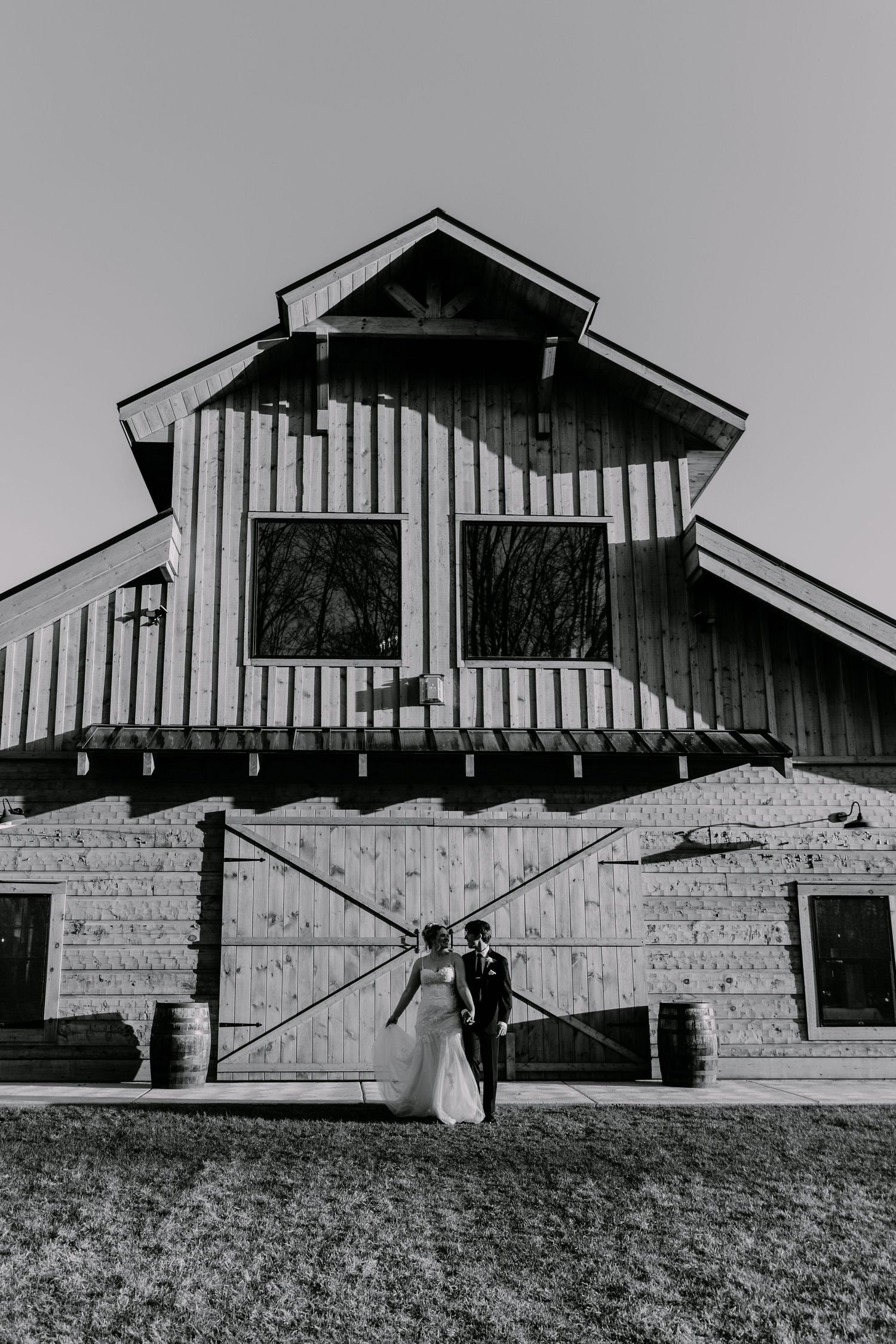 A wedding couple poses in front of a rustic barn with vertical siding and stone base in black and white.